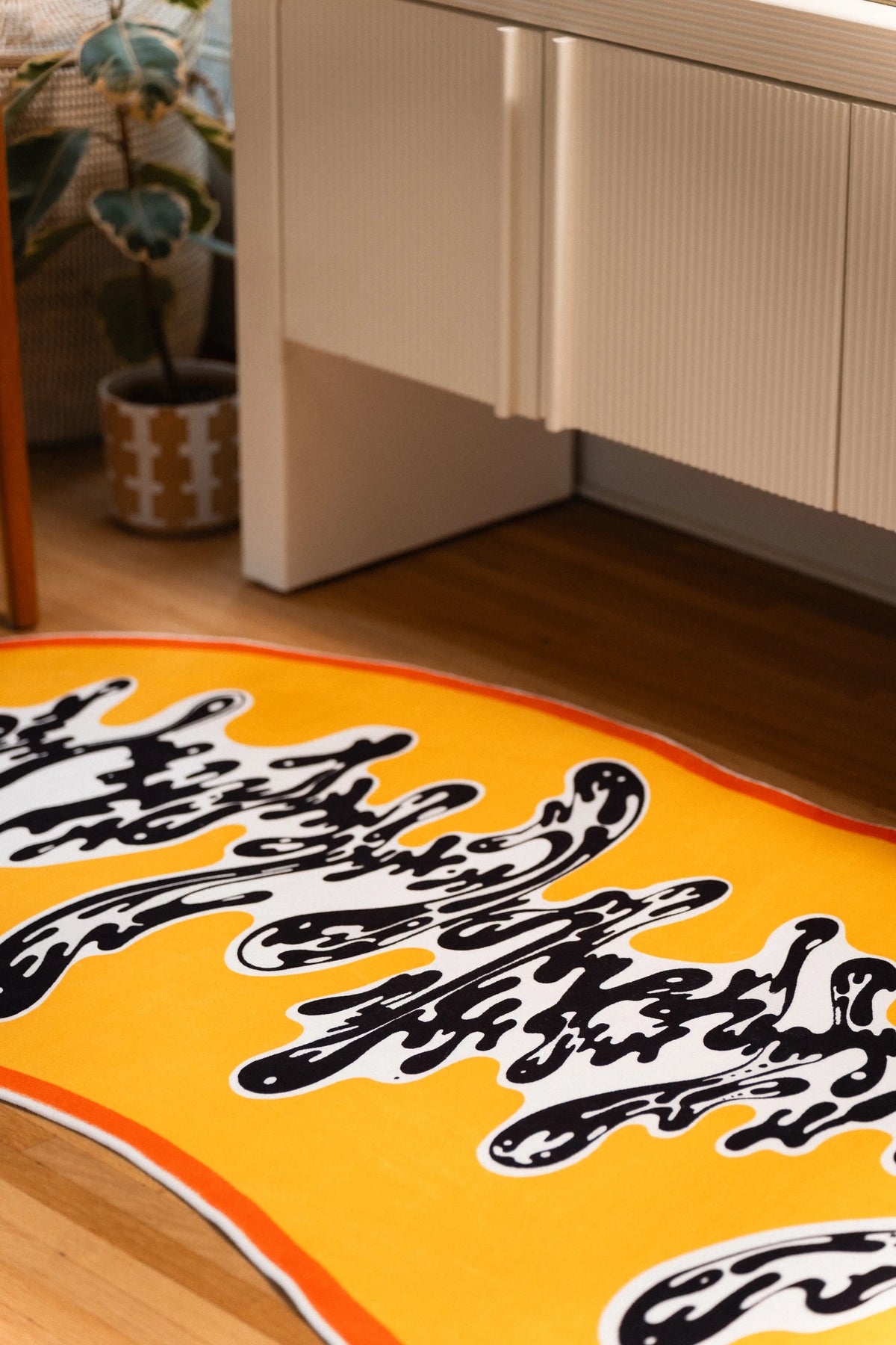 Ink-like black curls scatter across the white center of a golden yellow rug beneath a white cabinet and potted plant.