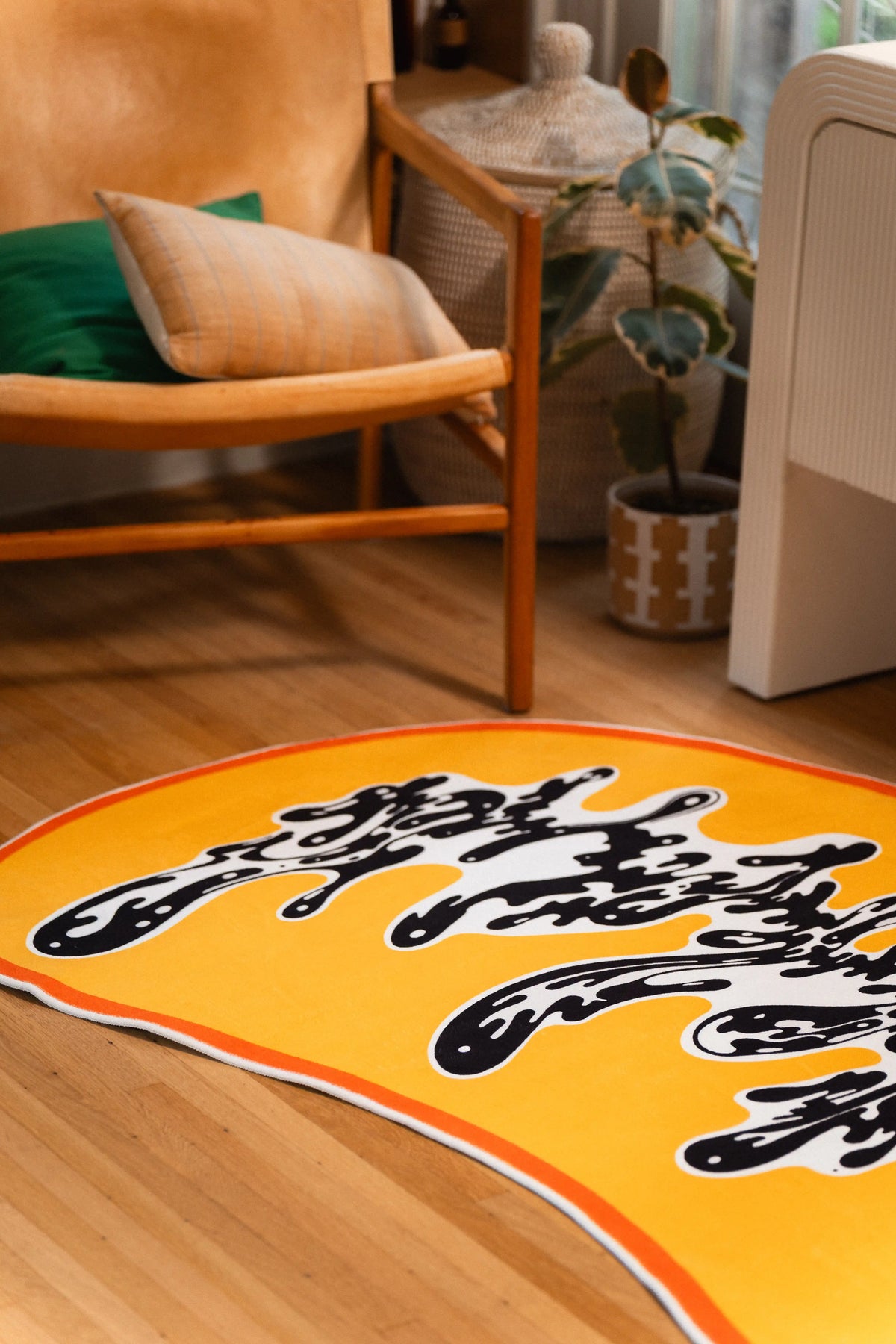 The orange-edged rug reaches toward a wooden lounge chair, with loose black strokes spreading across its yellow field.