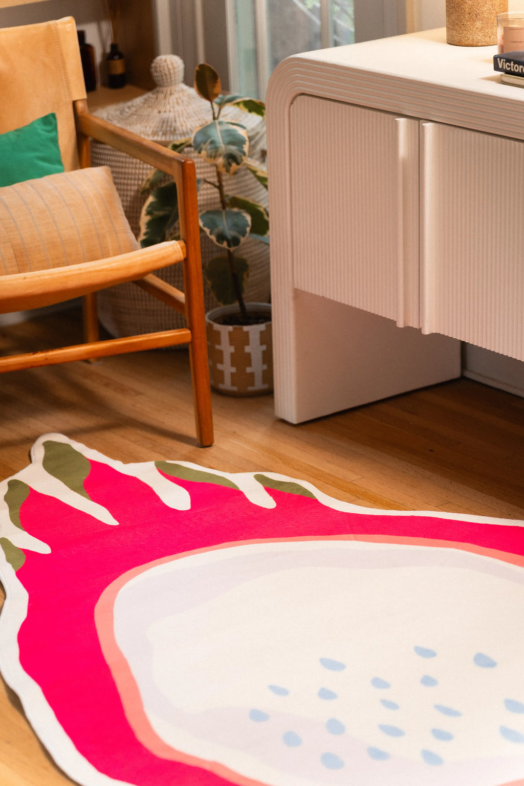 Hot pink and pale dragon fruit rug beside a wooden chair, potted plant, and white cabinet.