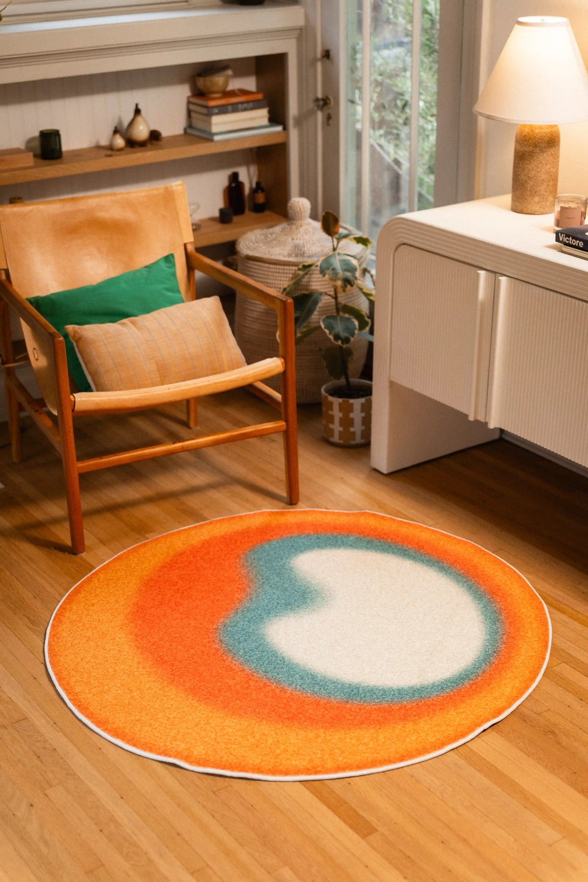 4' × 4' round rug with an orange outer ring, teal inner ring, and off-white center in front of a wooden armchair.