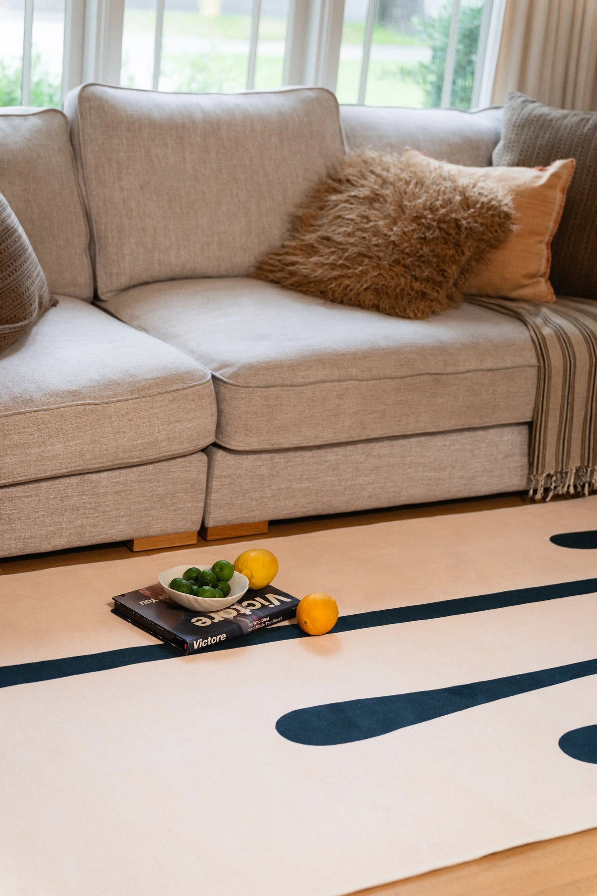 Beige area rug with green melting lines in front of a gray sofa, with a tray and fruit on top.