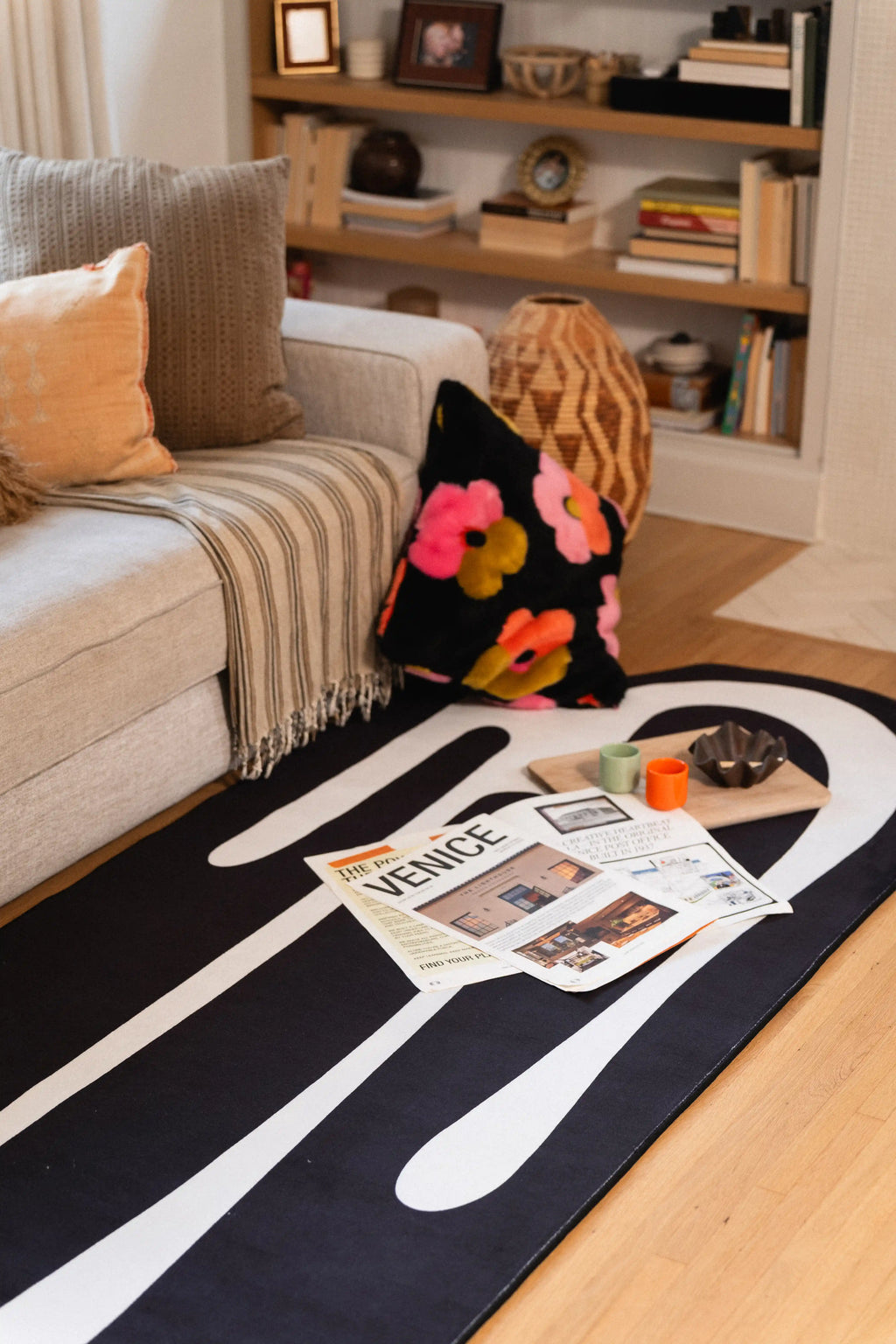 Black and white abstract rug beside a gray sofa with newspapers, a candle, and a floral cushion.