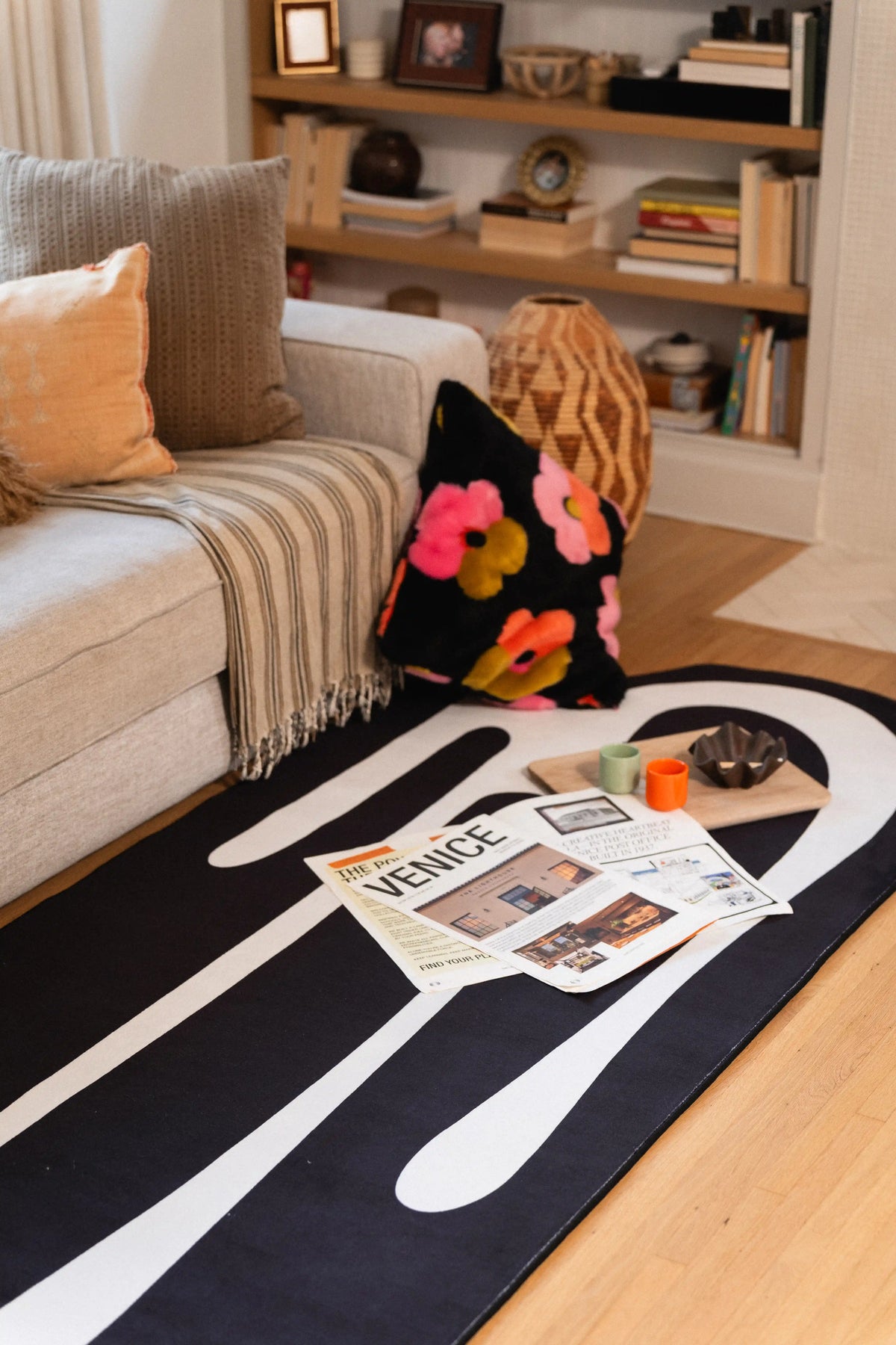 Black and white abstract rug beside a gray sofa with newspapers, a candle, and a floral cushion.