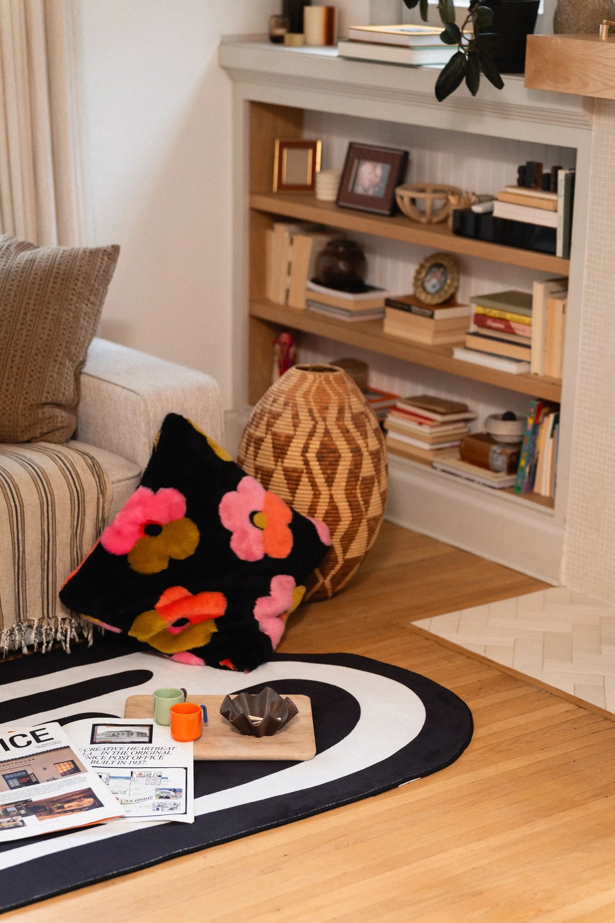 Black drip-pattern rug beside a gray sofa and floral cushion, with newspapers and small décor on top.