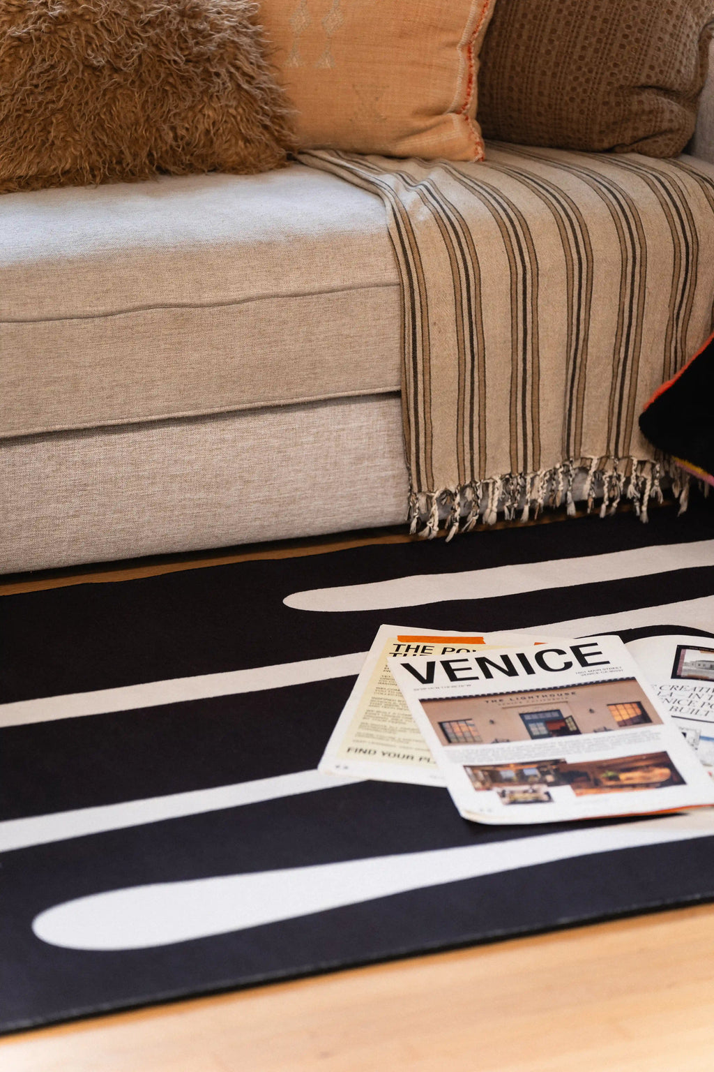 Black rug with flowing white lines beneath a gray sofa and striped throw, with a newspaper resting on top.