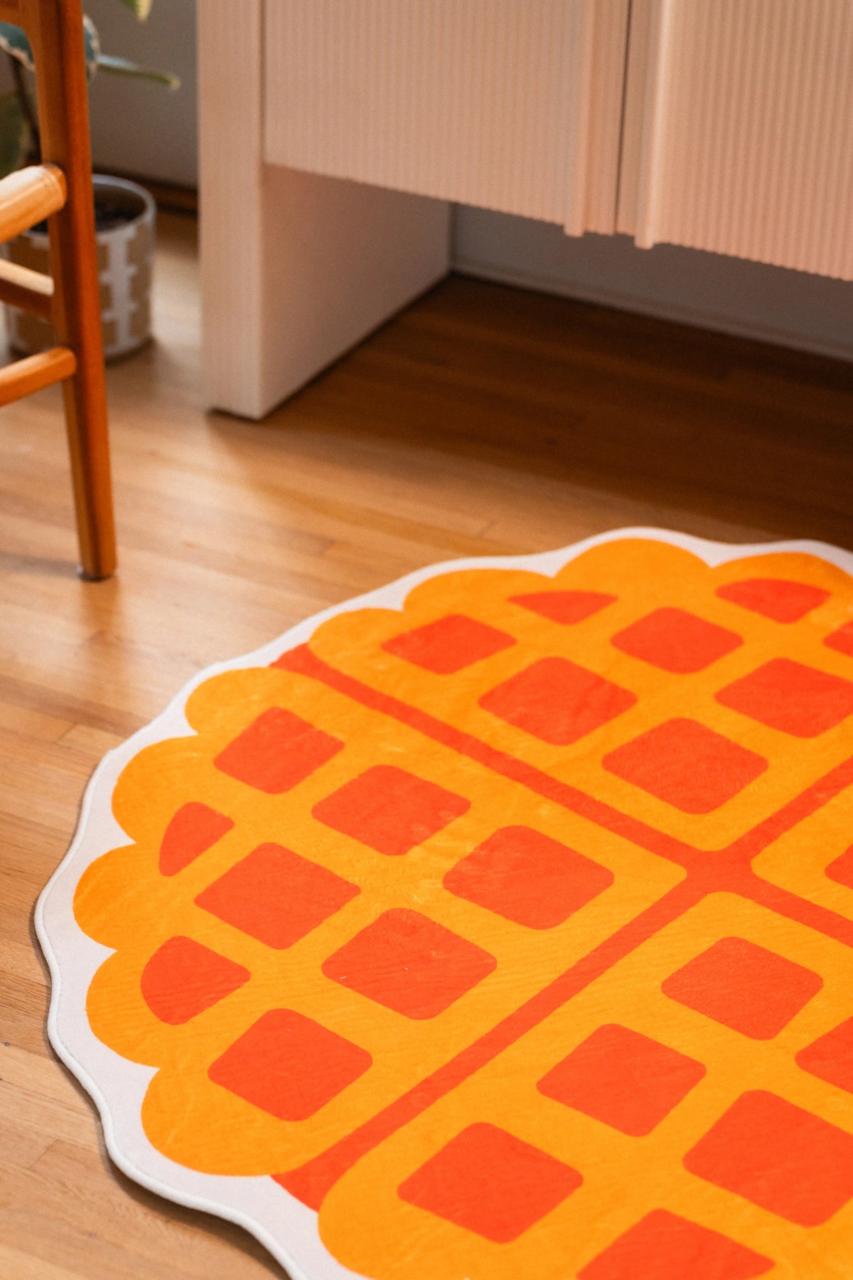 Orange grid squares on a yellow waffle rug with a cream scalloped edge beside a white cabinet and chair leg.