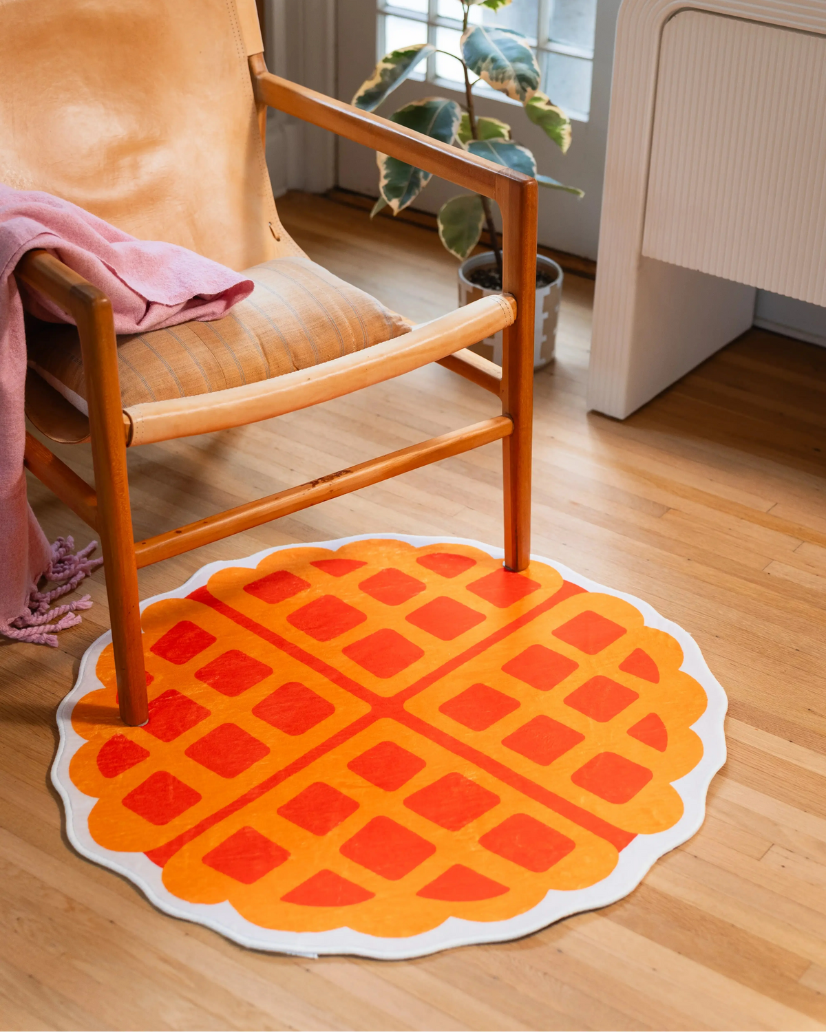 Round yellow waffle rug with an orange grid pattern and cream edge beneath a wooden chair beside a potted plant.
