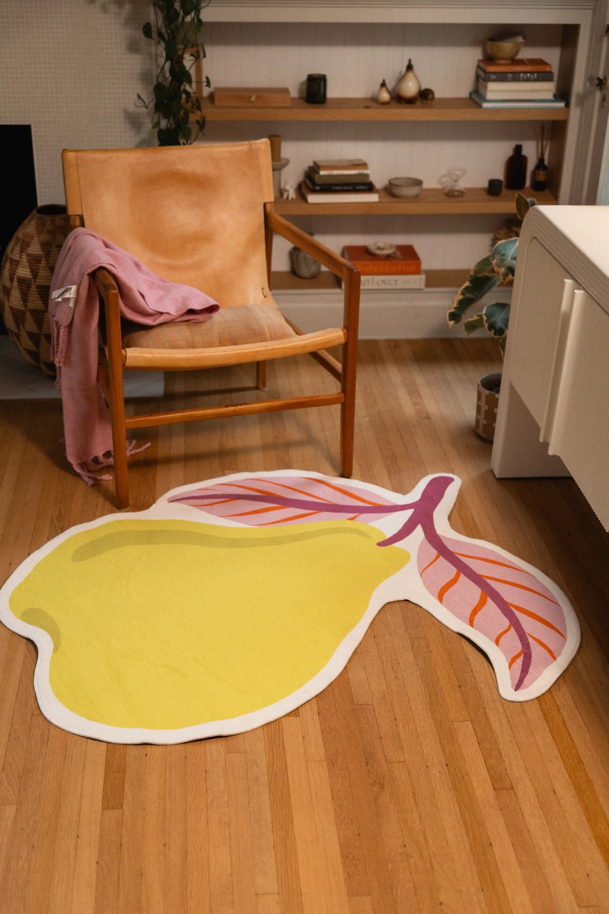 Yellow pear rug with a mauve stem, orange-veined pink leaves, and white outline in front of a wooden chair.
