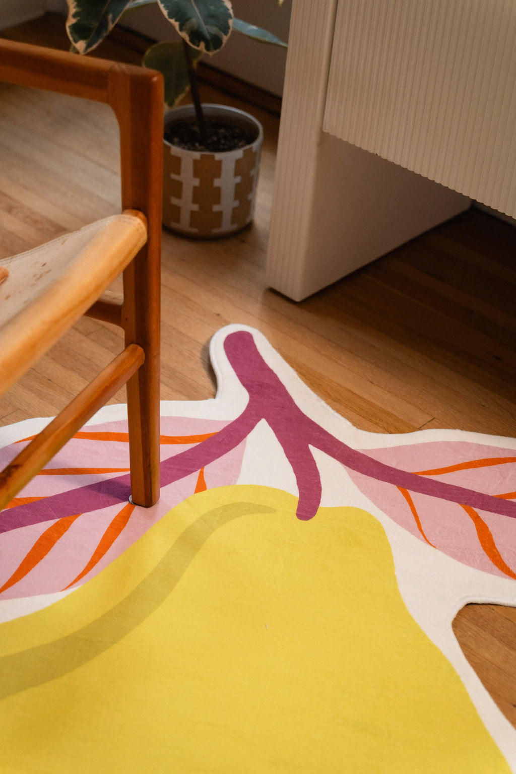 Mauve stem and orange-veined pink leaves on a yellow pear rug beside a wooden chair, potted plant, and white cabinet.