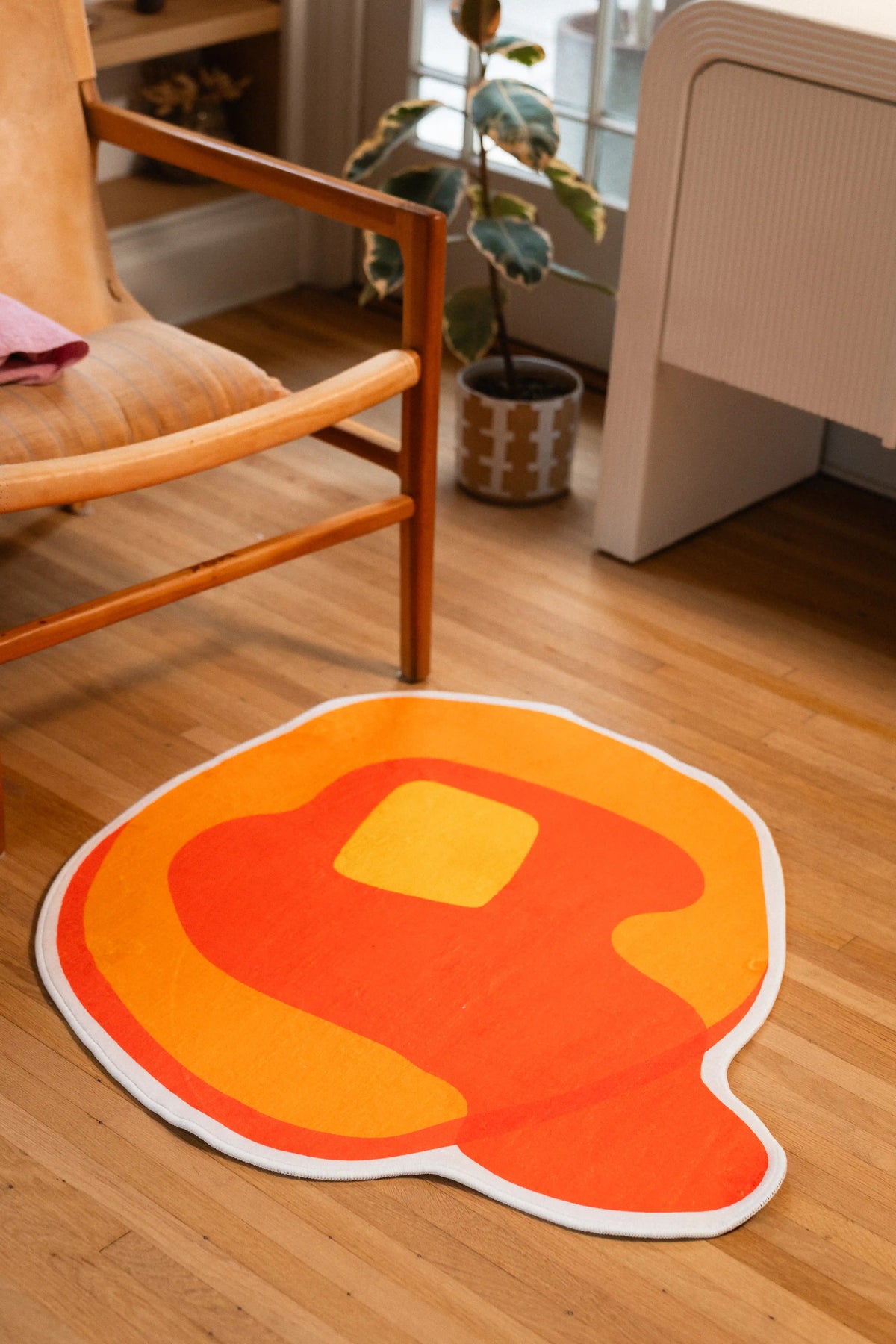 Yellow pancake rug with an orange syrup drip and square butter pat beside a wooden chair and potted plant.