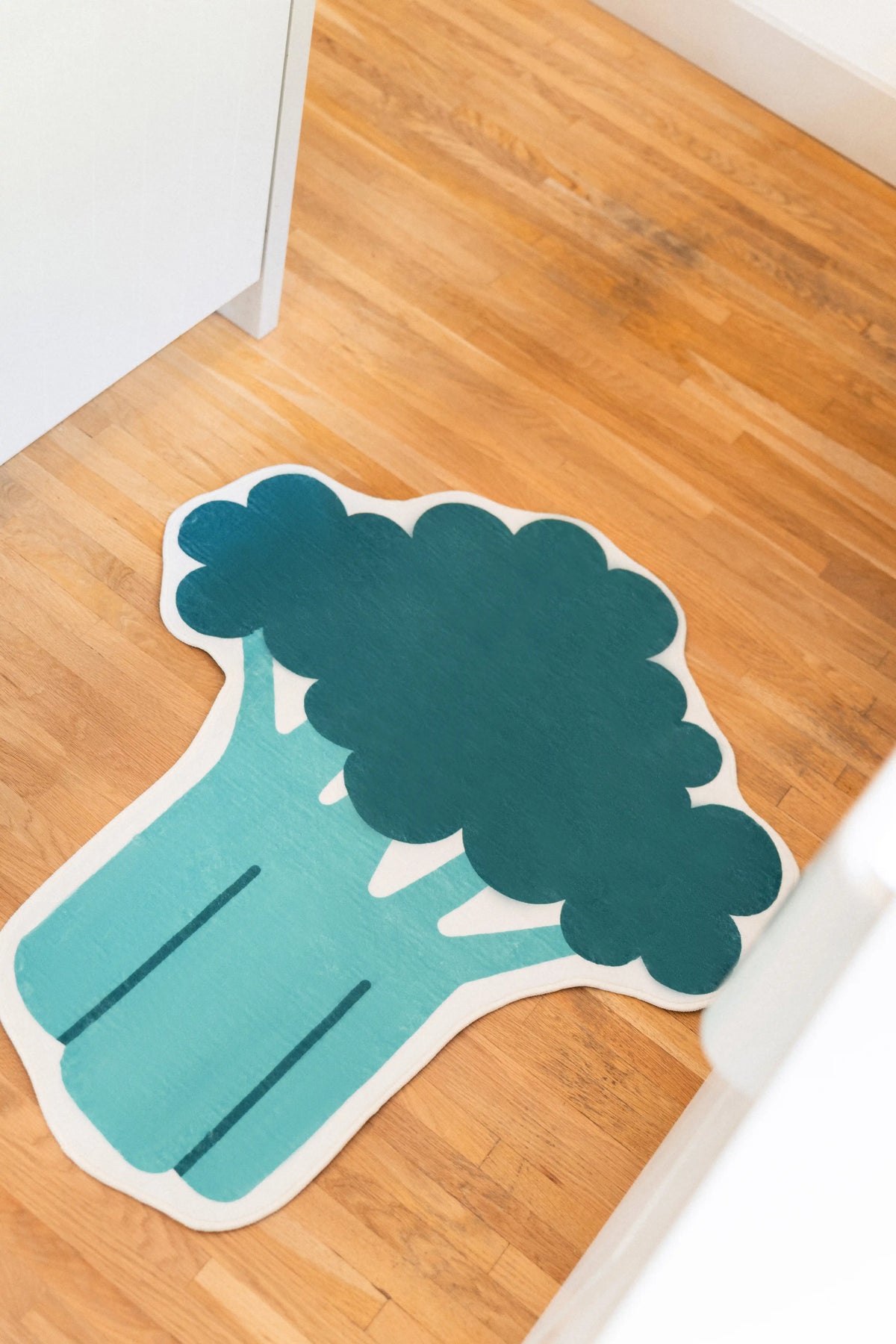 Green broccoli rug with rounded florets, a long teal stem, and white outline on a wood floor between white cabinets.
