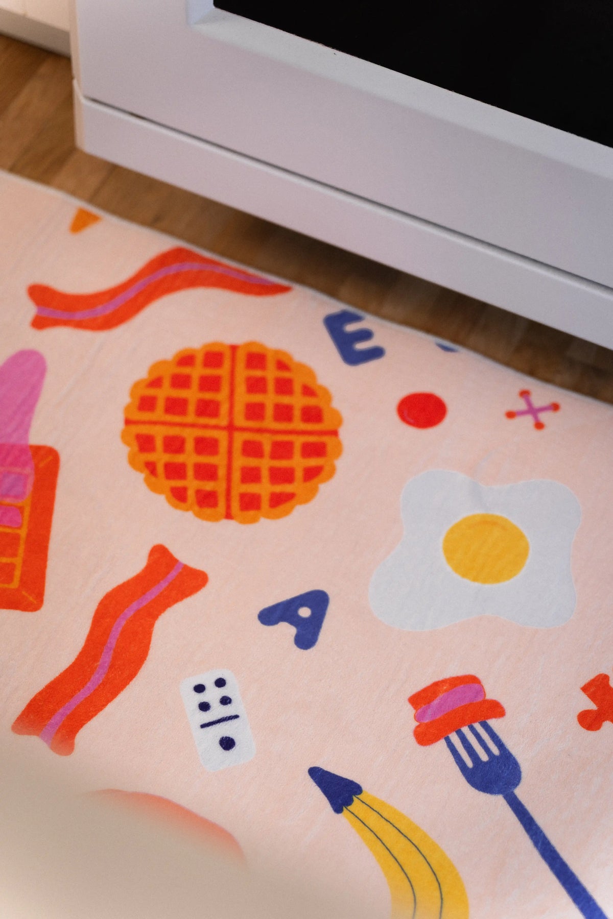 Orange waffle, fried egg, bacon strips, blue fork, banana, letters, and dots on a peach rug beneath a white oven.
