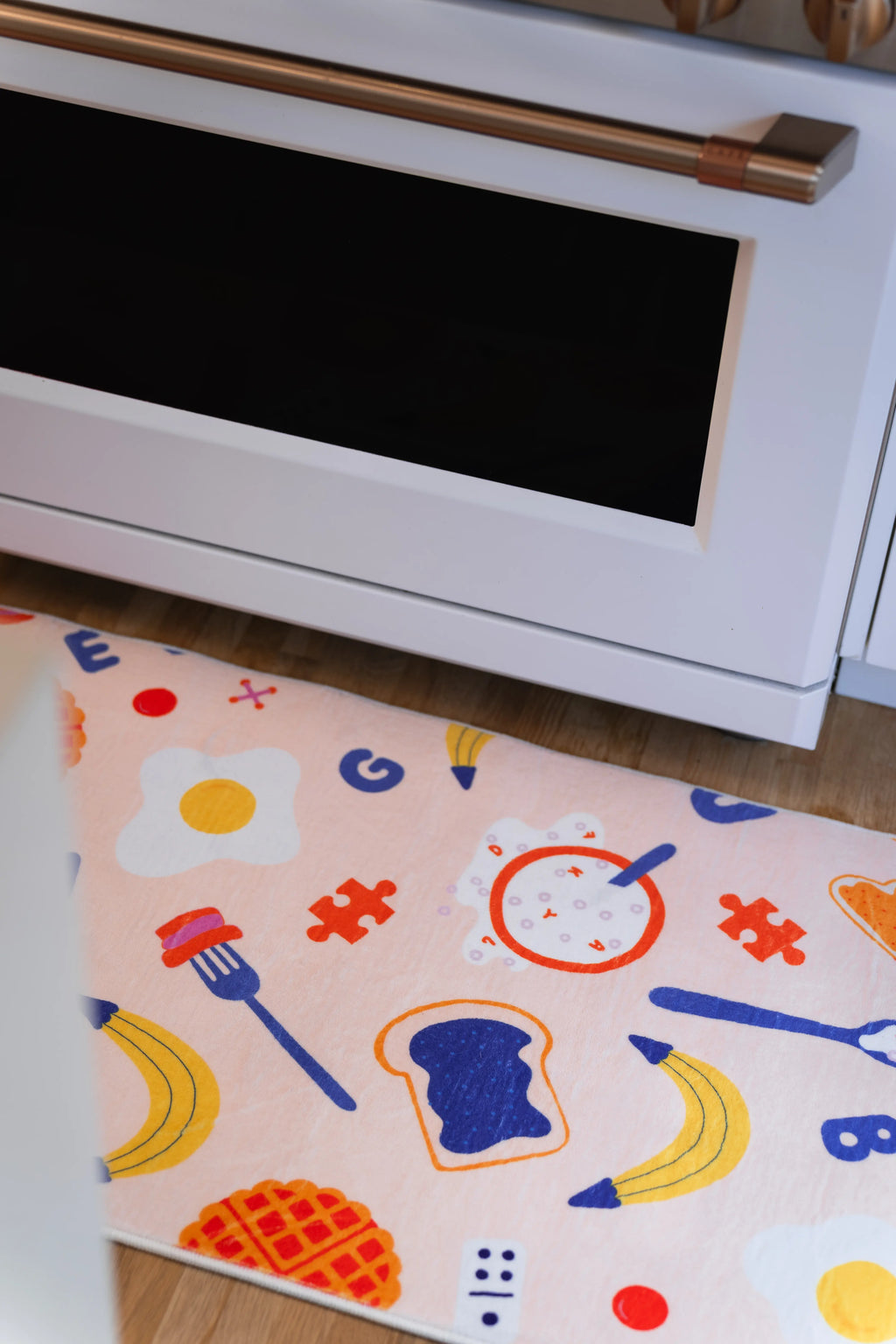 Fried egg, clock, blue toast slice, bananas, fork, waffle, and red puzzle pieces on a peach runner in front of a white oven.