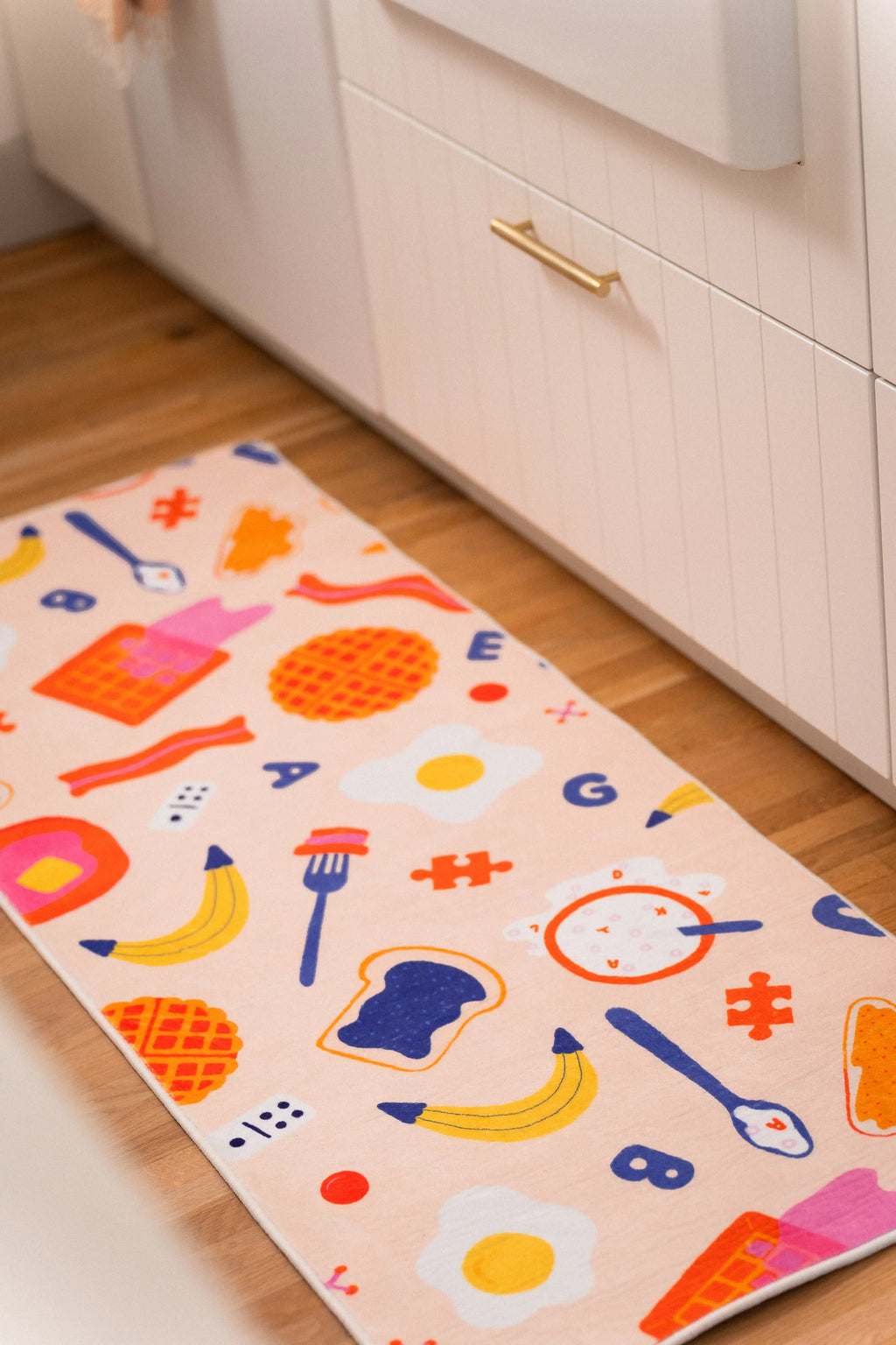 Peach kitchen runner with waffles, fried eggs, bacon, bananas, toast, utensils, letters, and red symbols beside white cabinets.
