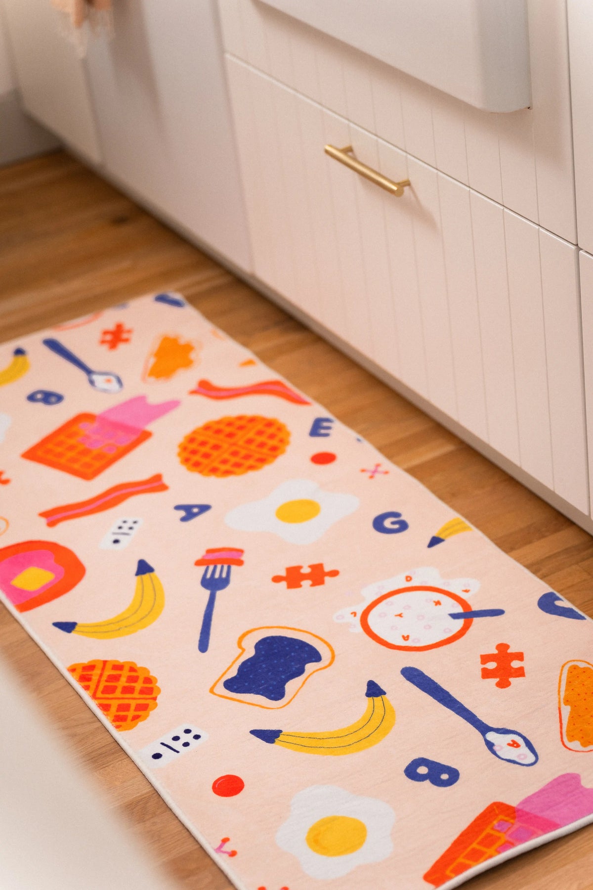 Peach kitchen runner with waffles, fried eggs, bacon, bananas, toast, utensils, letters, and red symbols beside white cabinets.