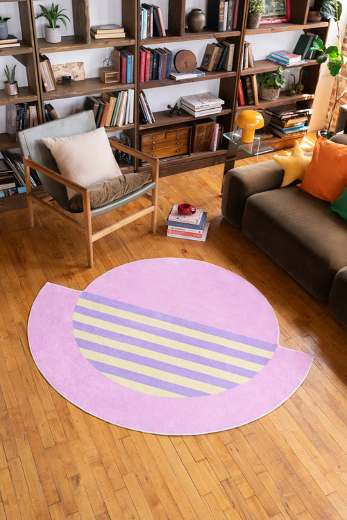 A modern area rug in pink and lilac with striped accents placed in a cozy reading nook with bookshelves