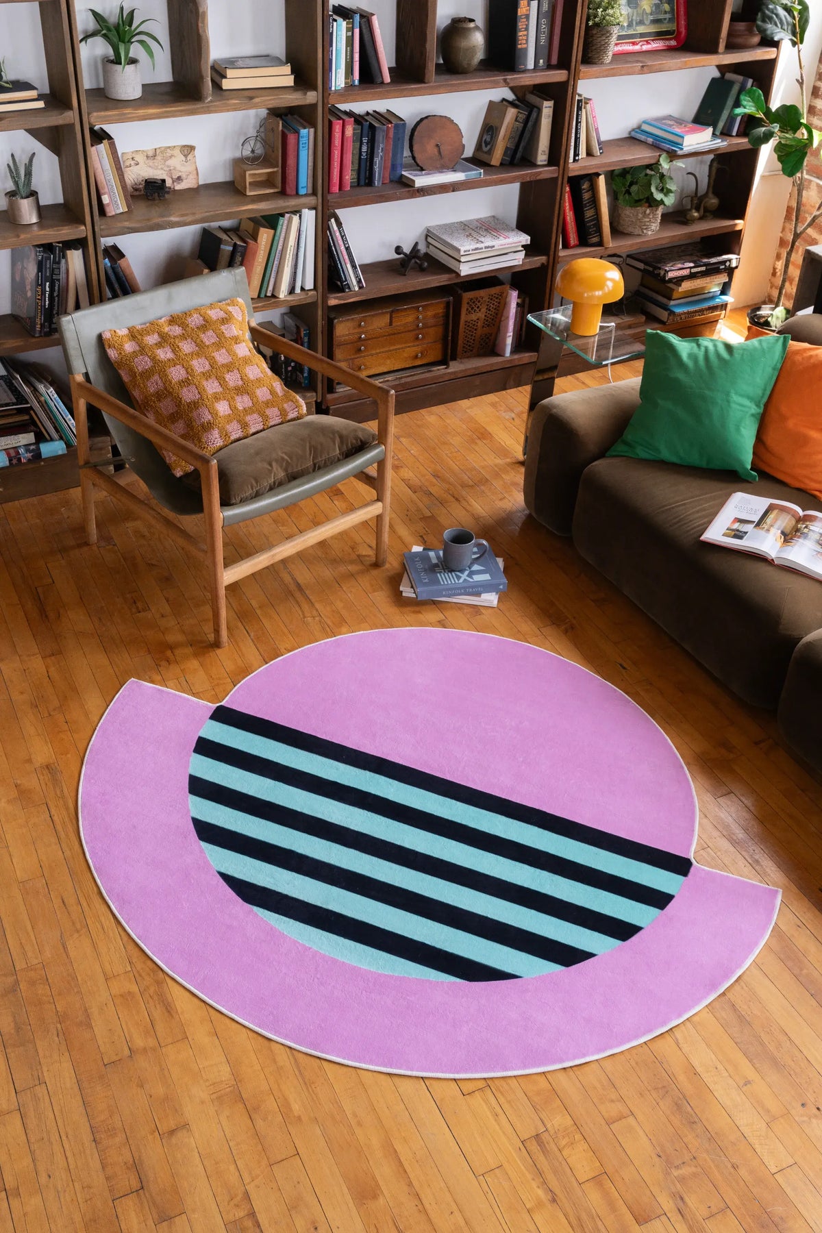 Modern abstract circle rug in pink and teal with black stripes, styled in a cozy reading nook with bookshelves and armchair