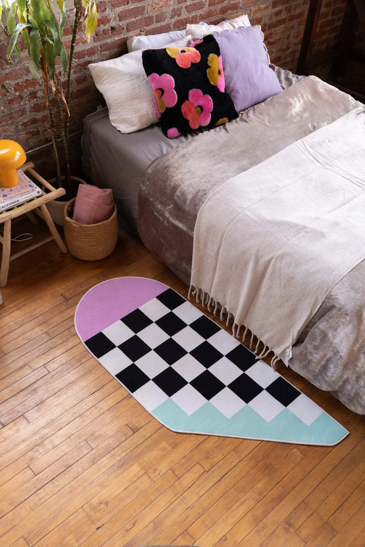 Pink and Blue Asymmetrical Checker Rug by Jakob Mizrahi placed beside a low bed in a bedroom with wood floor and brick wall