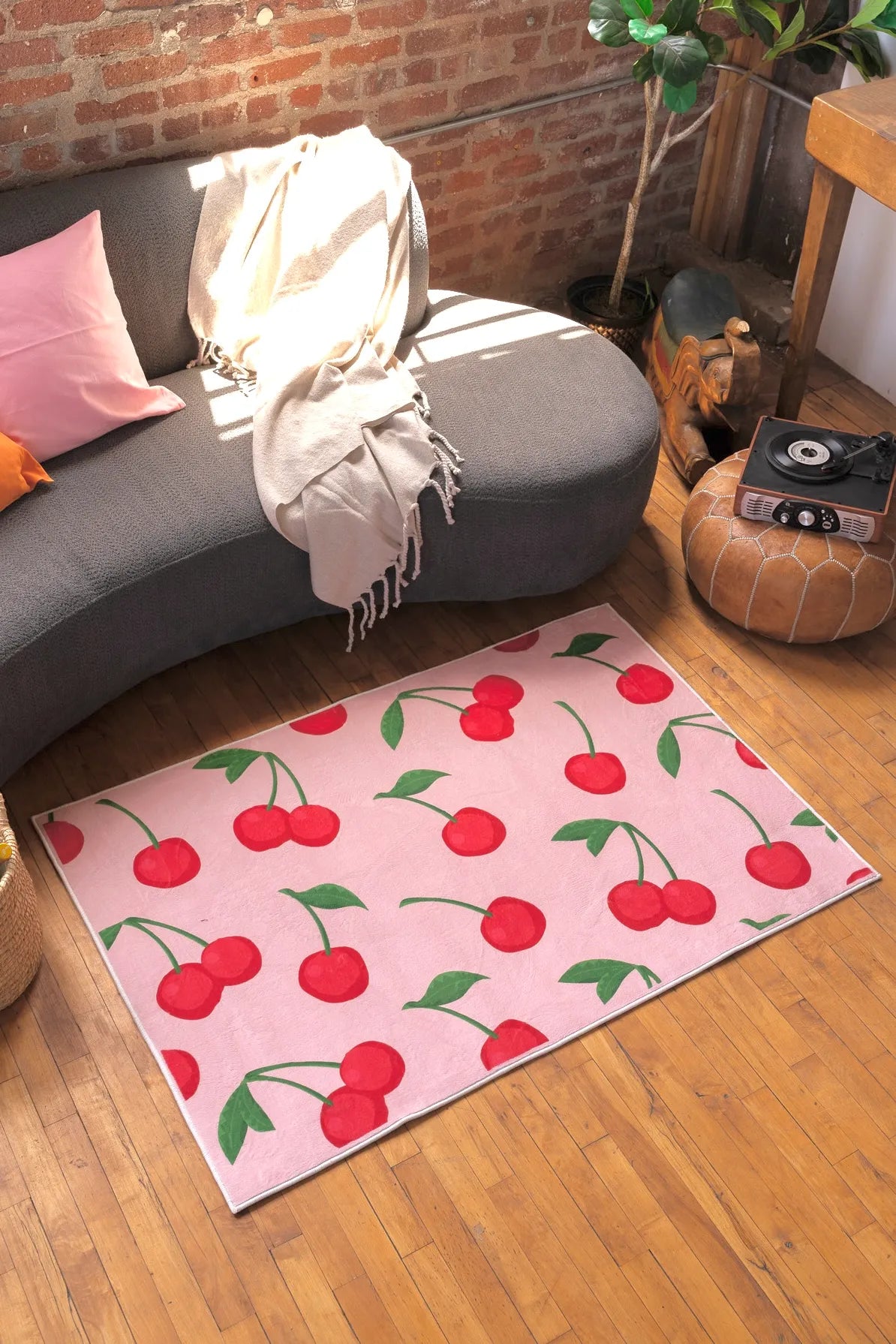 Pink rug with red cherry pattern and green leaves, styled beside a gray sofa and round pouf in a sunlit corner.