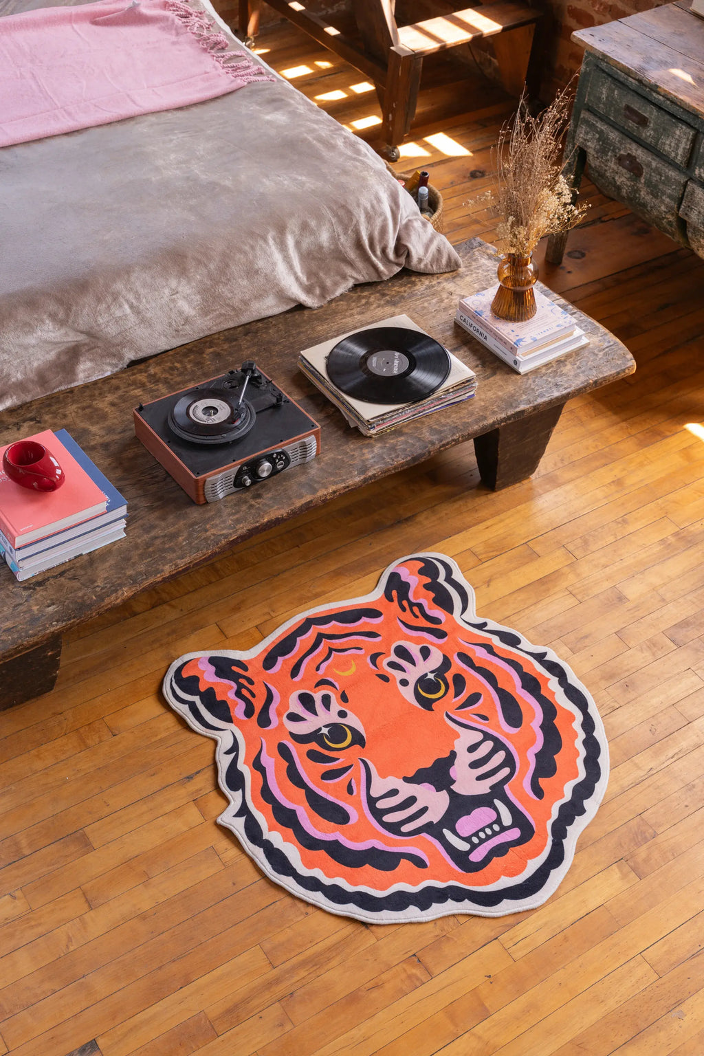 Funky tiger rug by a rustic low table and bed, on warm-toned wood floor in a sunlit loft-style bedroom.