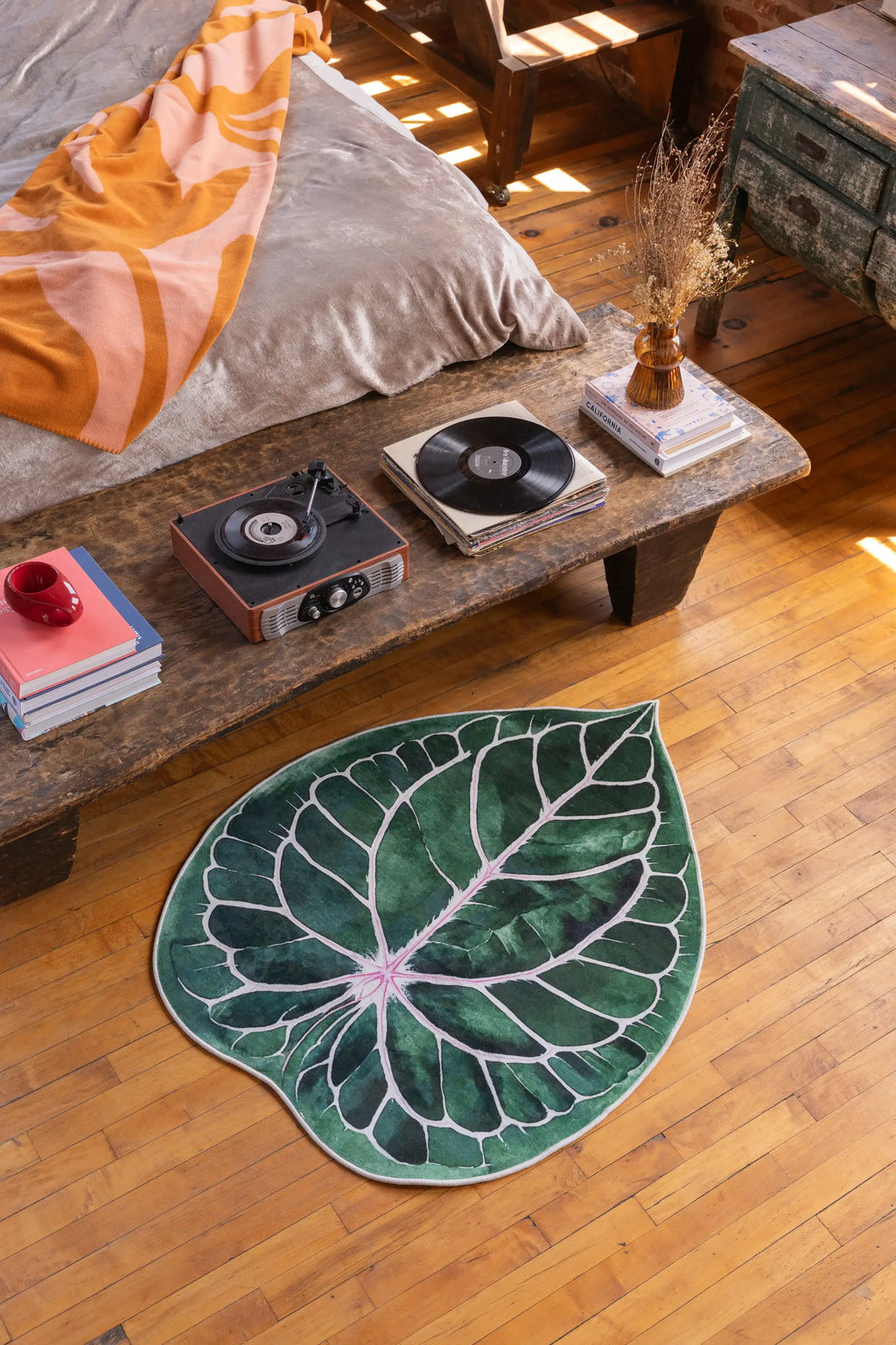 Irregular Caladium Leaf Rug by Johann Banta placed on wood floor beside a rustic table, near a bed in a cozy corner.