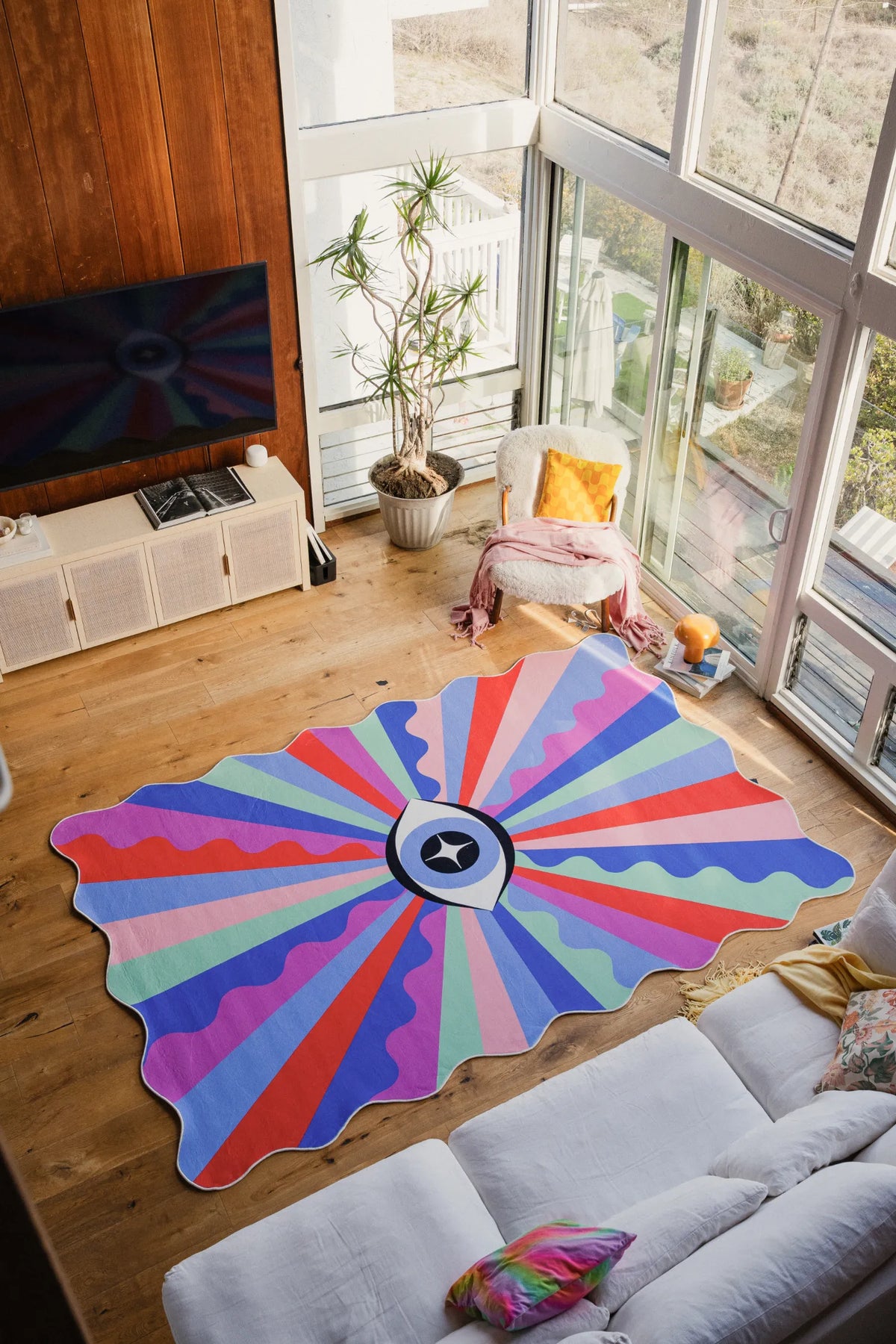 Colorful Lazer Rays Eye Rug by Jessica Miller, 7' × 10', centered in an airy corner with wood floor and large windows.