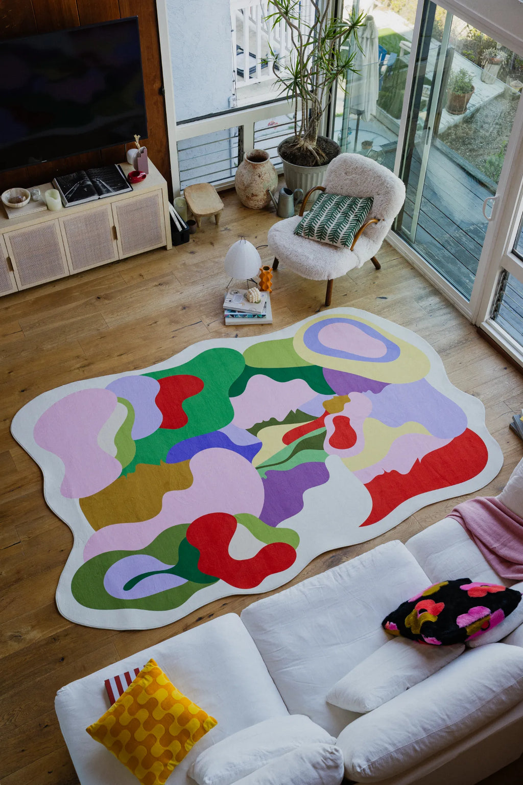 The pastel abstract rug with an irregular shape lies in a spacious, sunlit living room beside a white sofa, creating a vibrant centerpiece.