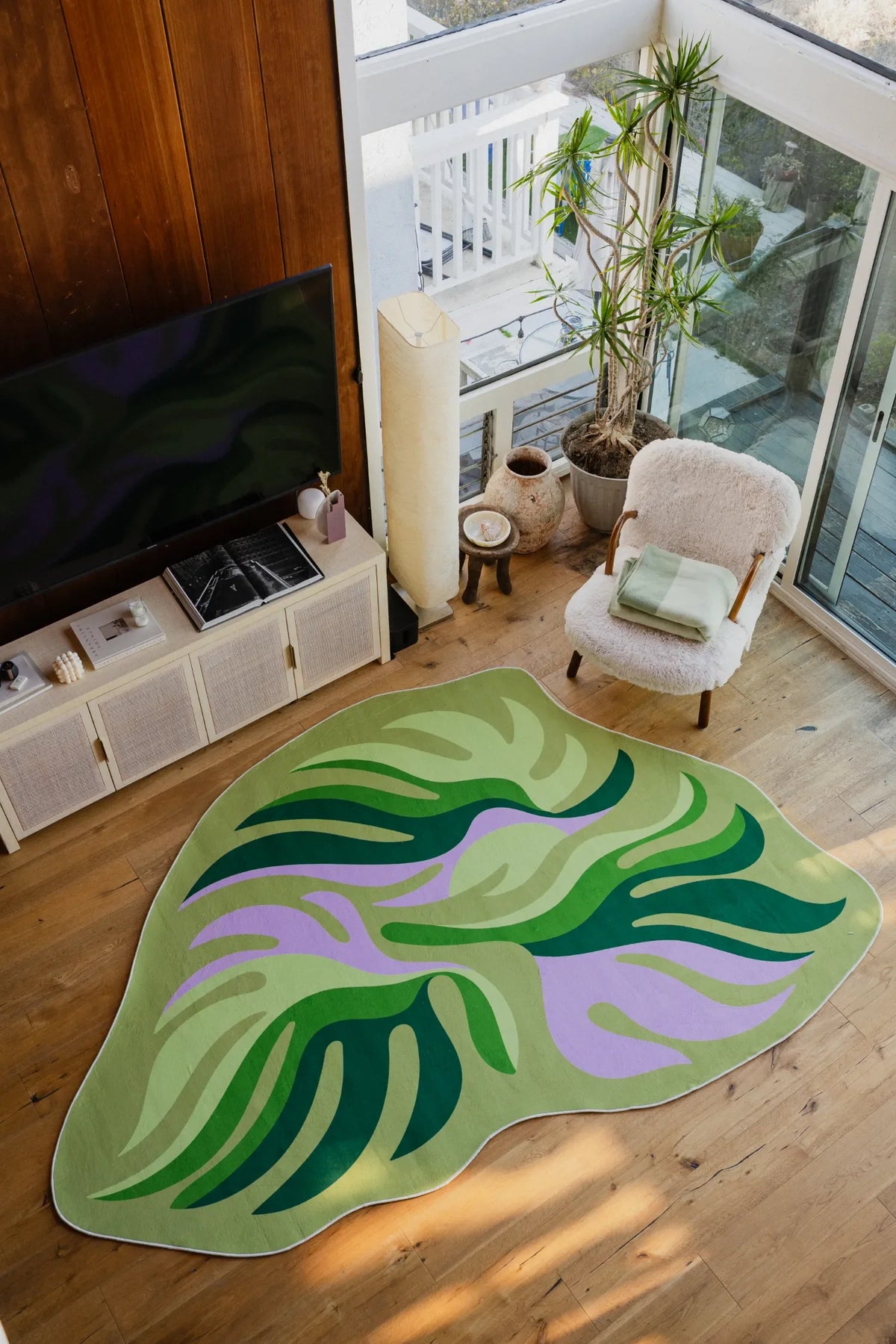 Leaf-shaped rug with green and lilac tones placed in front of a boucle chair and indoor plant by floor-to-ceiling windows.