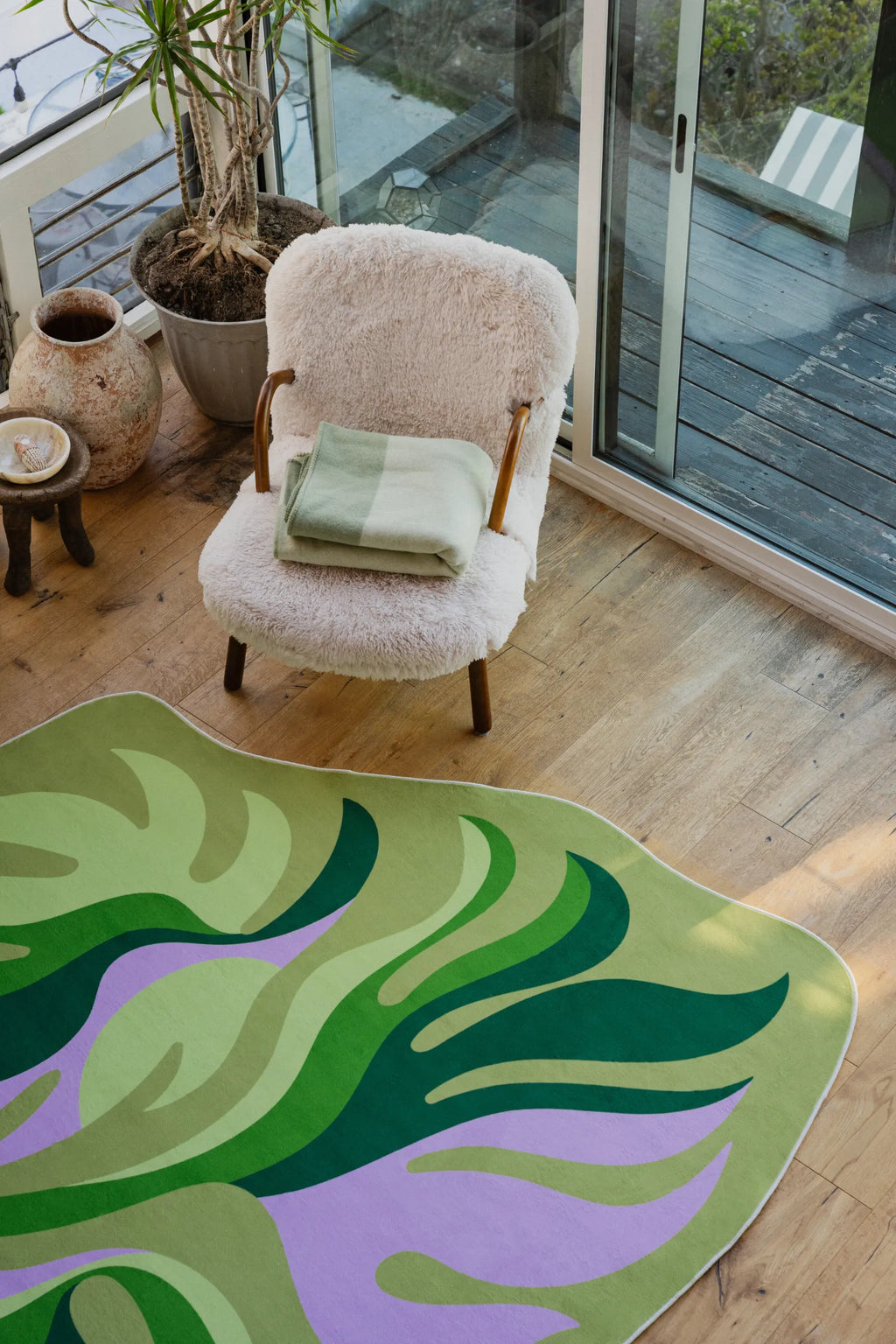 Botanical leaf-shaped rug styled with a cozy armchair and blanket, next to large windows in a sunlit corner.