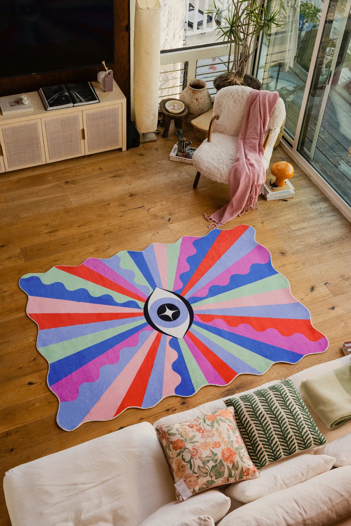 Colorful Lazer Rays Eye Rug by Jessica Miller, 5' × 7', laid on wood floor beside a soft armchair near full-height windows.