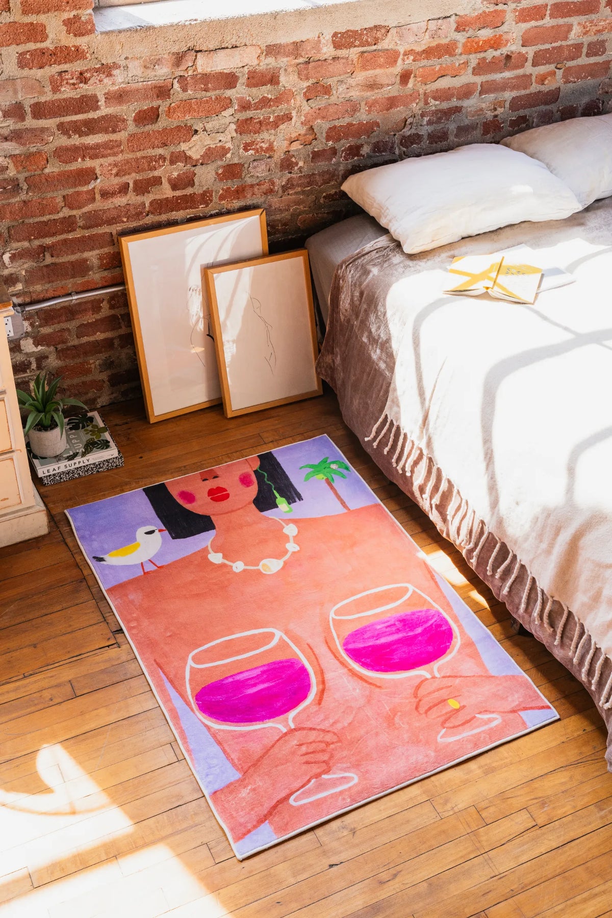 Medium 3'X4' woman art rug with wine design beside bed, side view showing brick wall, framed art and natural light streaming in.