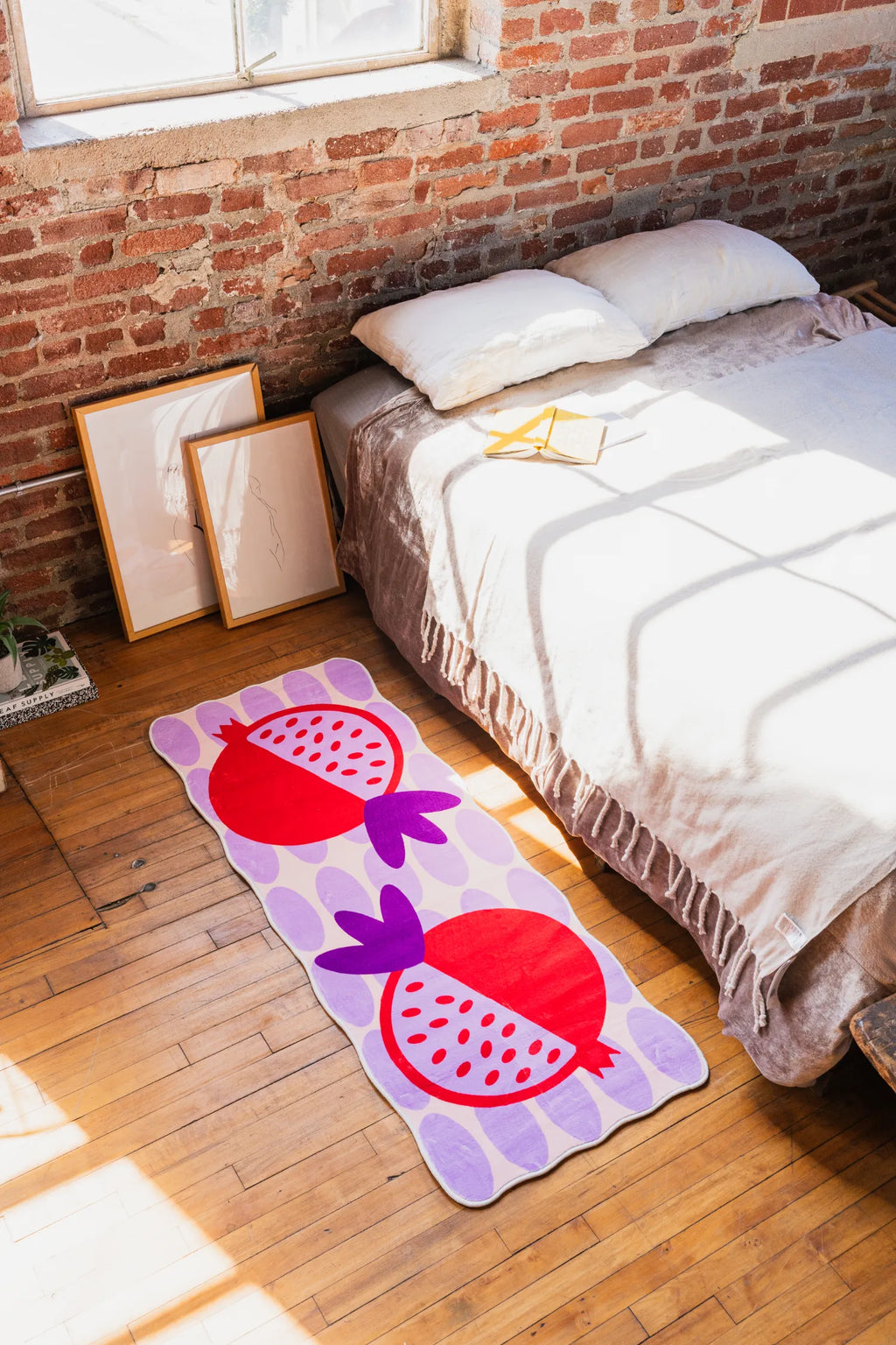 Red and Purple Pomegranate Fruit Area Rug by Shayla Goethe, styled beside a low bed in a loft bedroom with exposed brick walls and soft sunlight.
