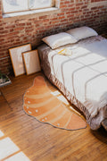 Buttery Orange Croissant Rug by Petra Lizde placed beside a bed on a wooden floor with sunlight streaming in