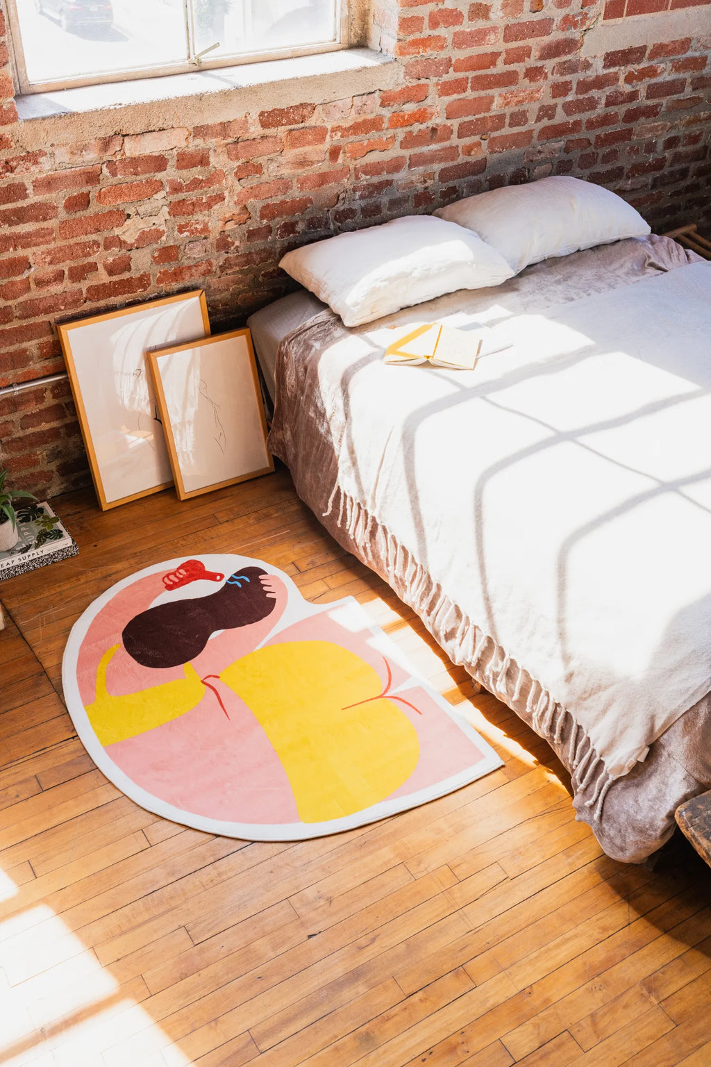 Funky Body Art Rug by Nimei in vibrant yellow and pink, styled beside a sunlit bed with exposed brick walls and leaning frames.