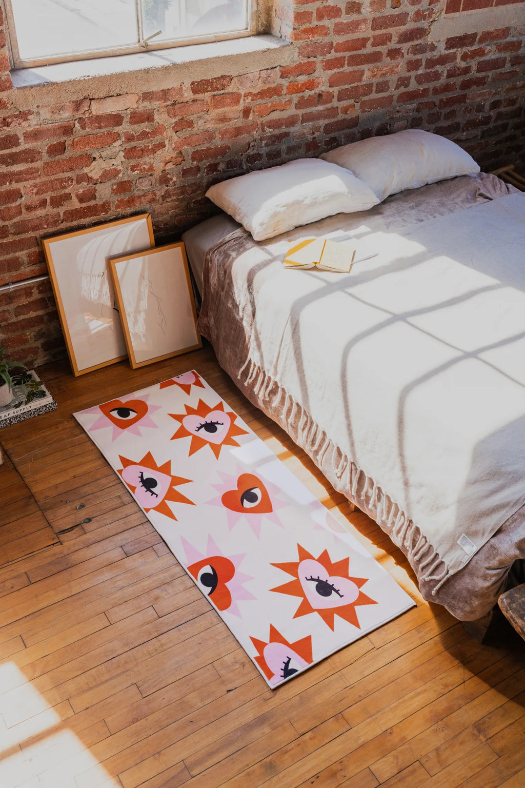 Playful rug with bold heart eye pattern placed beside low bed on wood floor near brick wall and leaning framed artwork