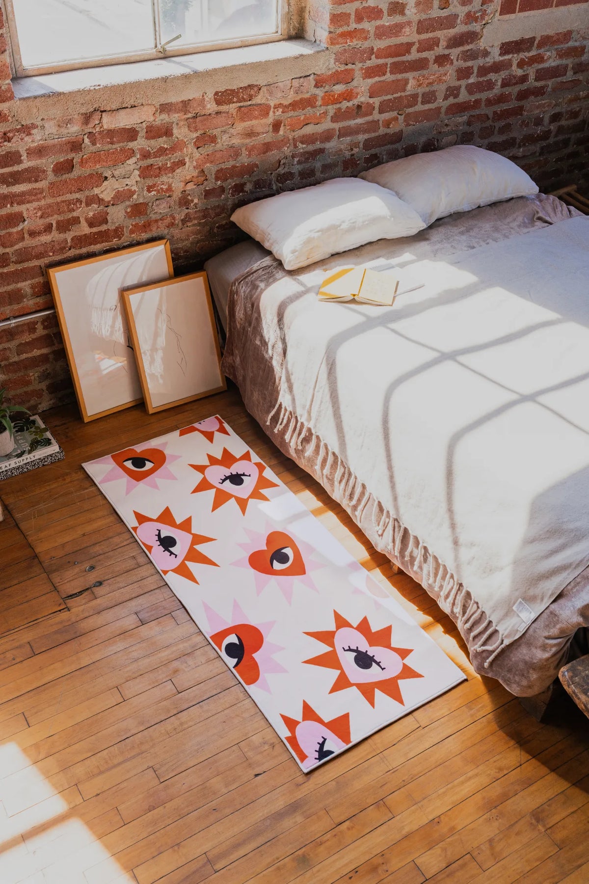 Playful rug with bold heart eye pattern placed beside low bed on wood floor near brick wall and leaning framed artwork