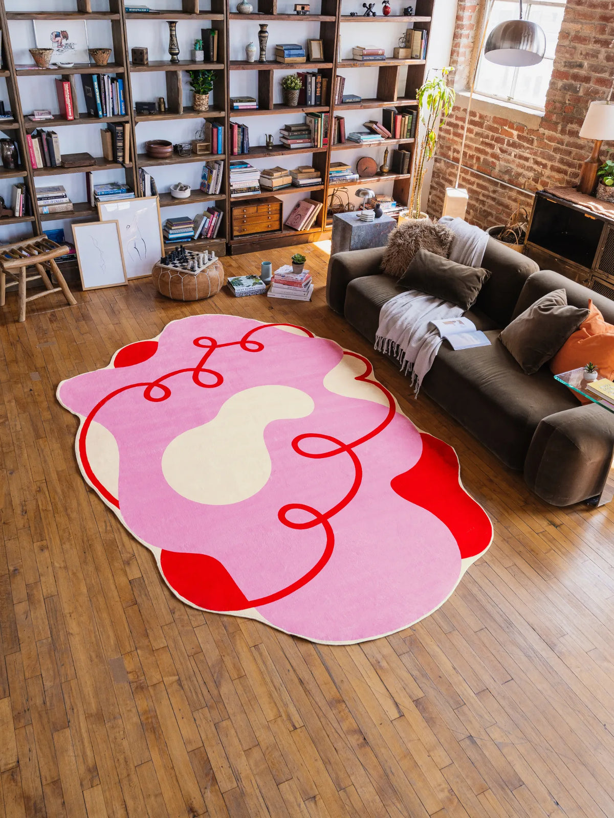 Wavy Abstract Irregular Area Rug in pink, laid out in a cozy living room with a brick wall, wooden floor, and a brown sofa filled with colorful pillows.
