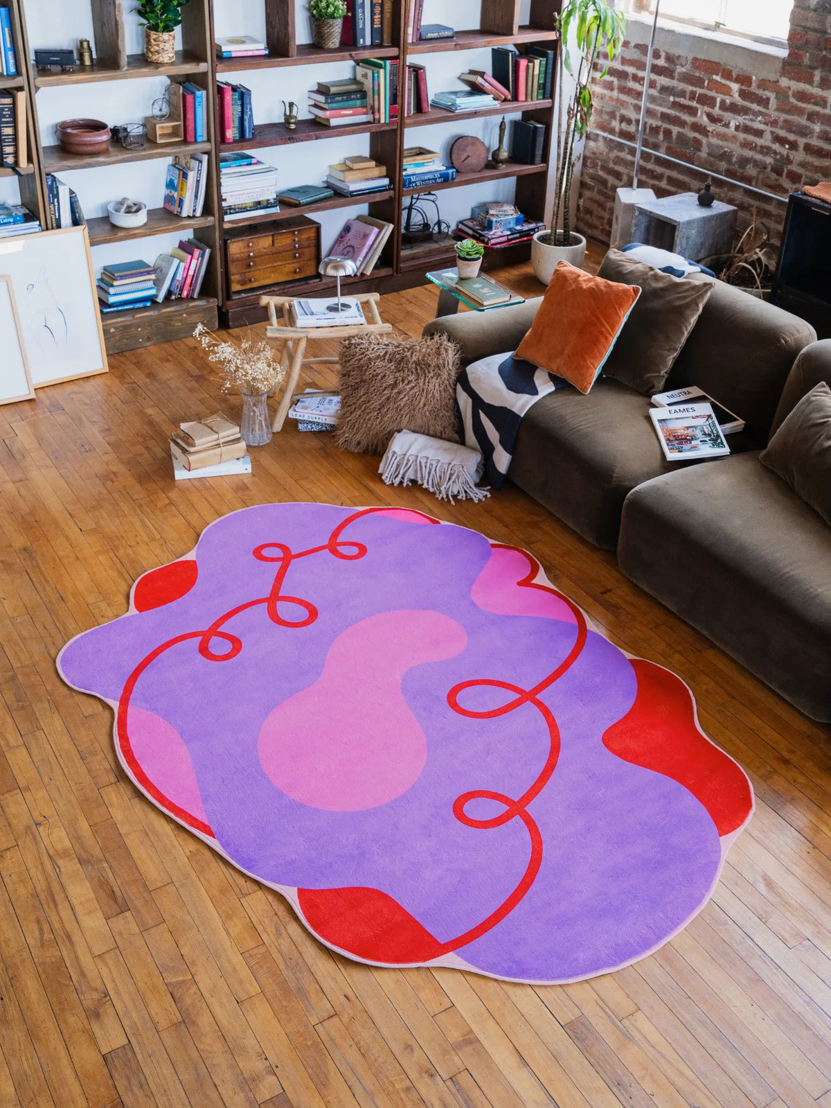 Playful irregular area rug in purple and pink with flowing red details, placed in front of a brown sofa and bookshelves for a modern artistic vibe.