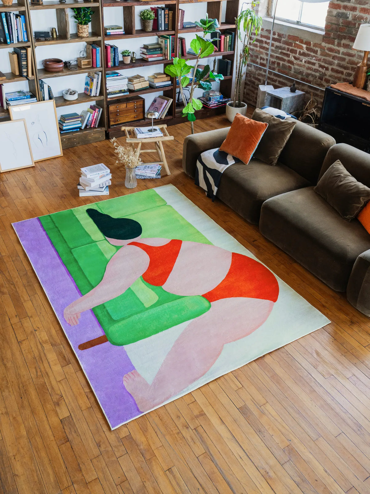 Medium 5'X7' abstract body rug with bold red and green forms, laid out near a sofa accented with orange pillows and framed artworks leaning casually on the wall.