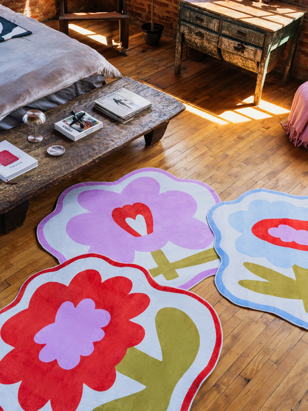 Three flower-shaped rugs in orange, pink, and blue arranged on wooden floor beside rustic bench in bedroom.