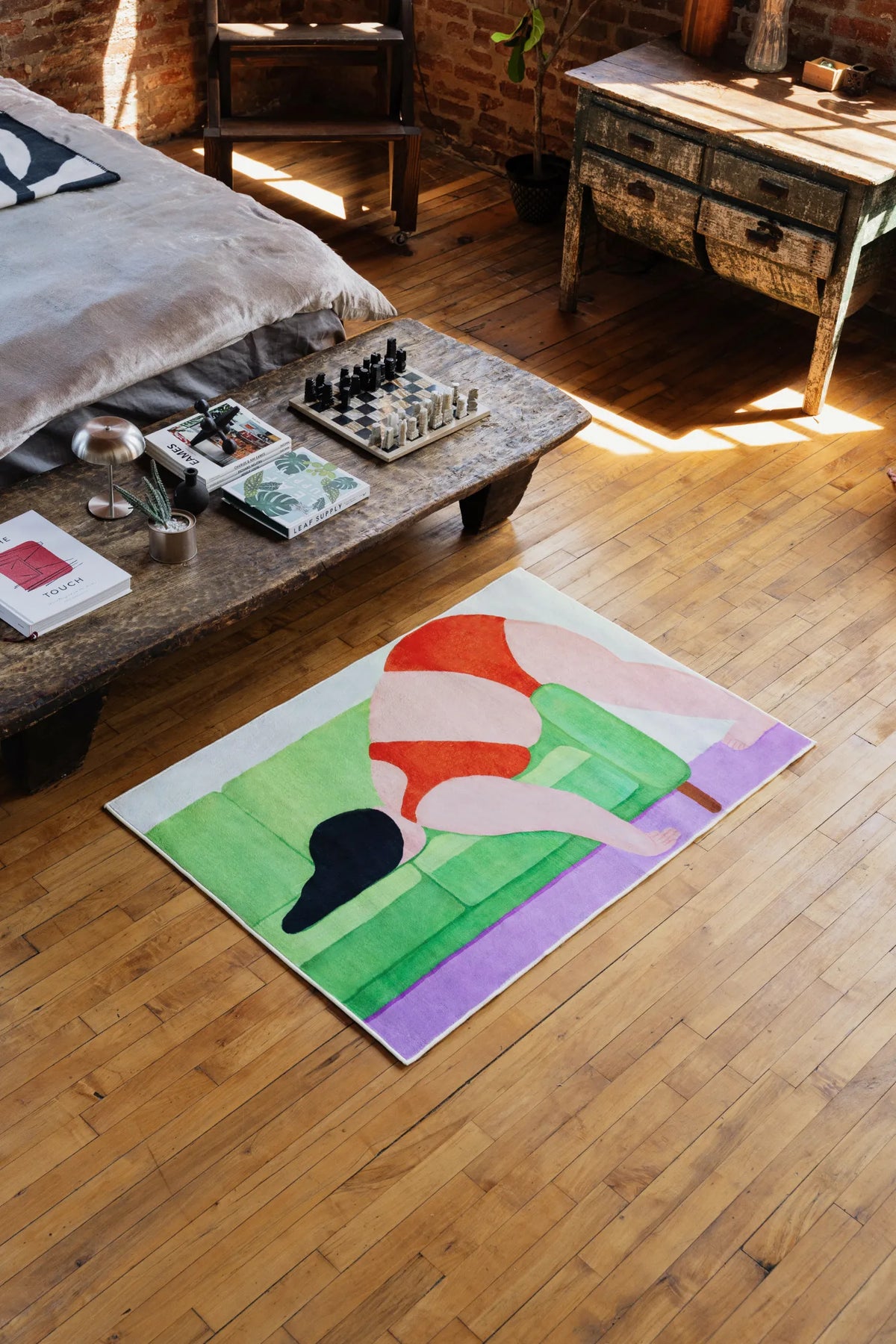 Compact 3'X4' body art rug in vivid colors, positioned at the foot of a rustic wooden bench beside a sunlit window, adding a playful accent to the bedroom.