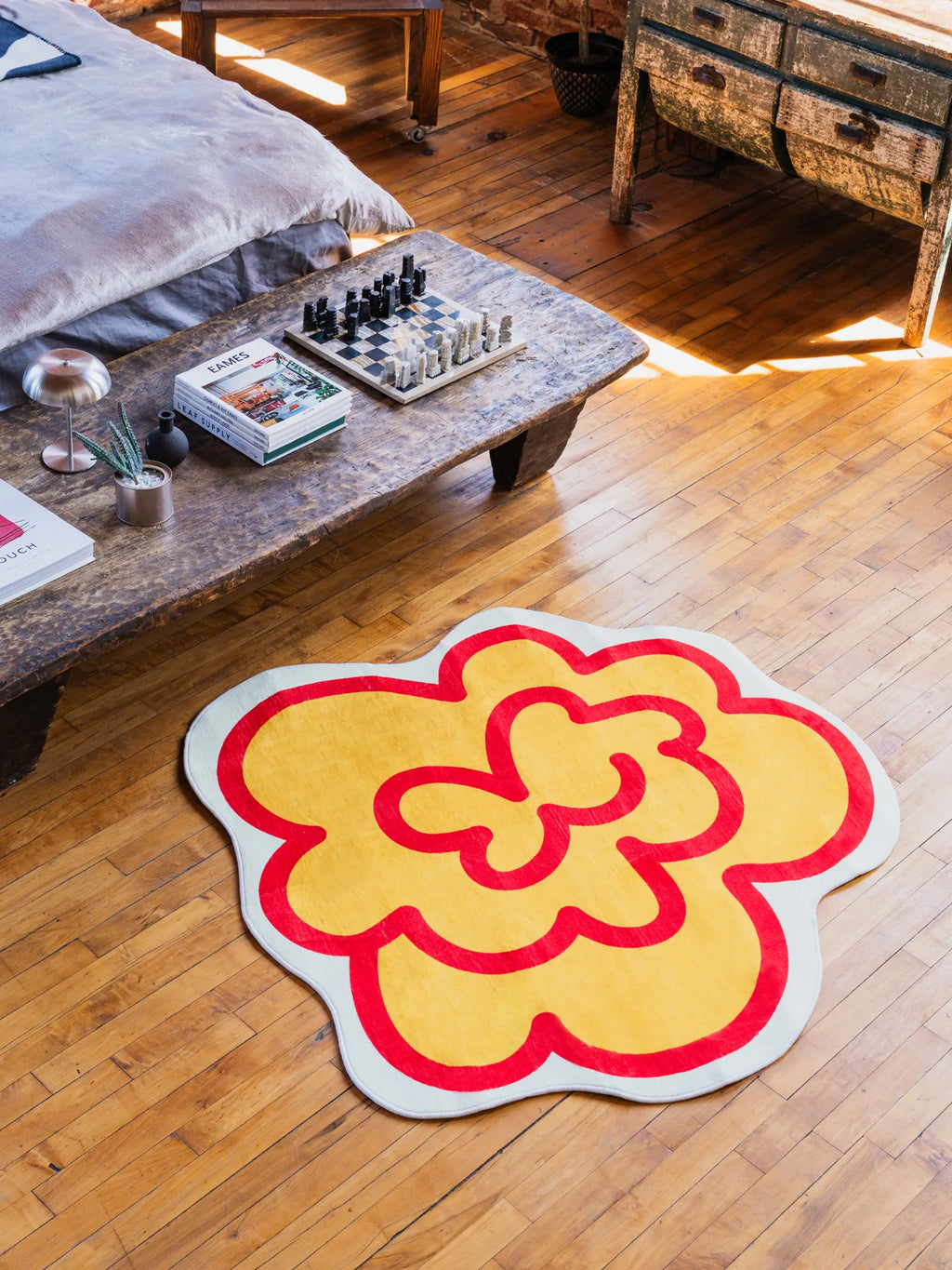 A yellow funky flower shaped rug rests on wood floor in a bedroom, adding playful contrast near the bed and rustic table.