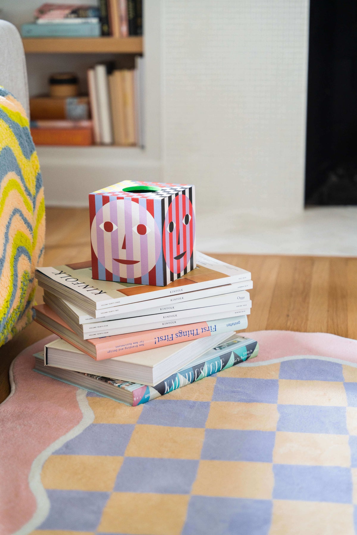 Close-up of the pastel rug’s scalloped pink border next to a playful stack of books and a striped face-print tissue box.