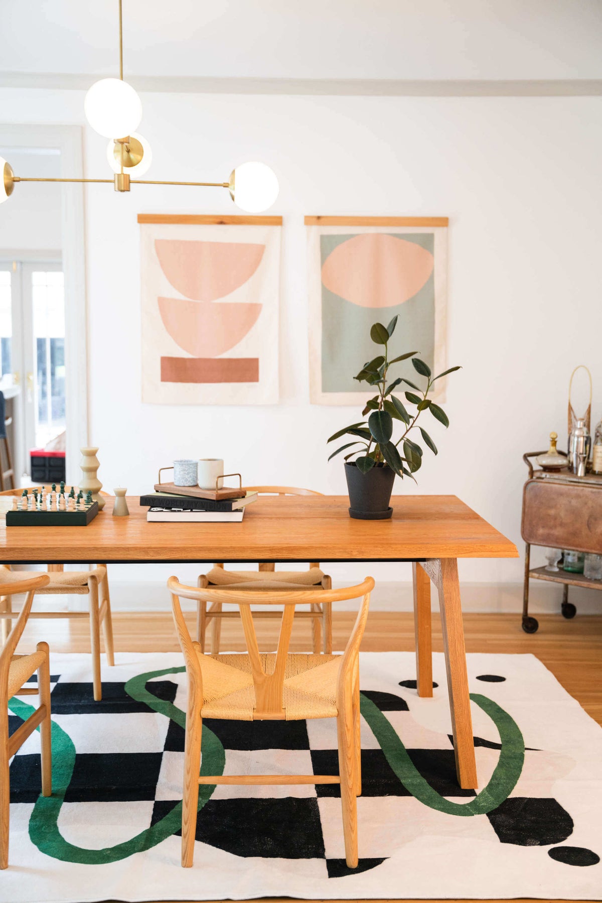 A black and white checkered rug with green lines brings bold contrast under a wooden dining table with woven chairs and pastel wall art.