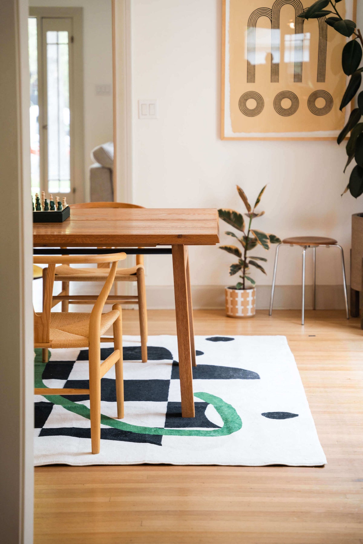 A minimalist dining space with a light wood table and chairs placed atop a black-and-white checkered rug with green curves.