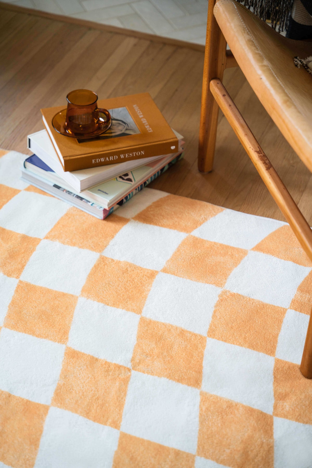 Corner of the wavy checkered rug styled with stacked books and an amber glass cup beside a tan leather chair.