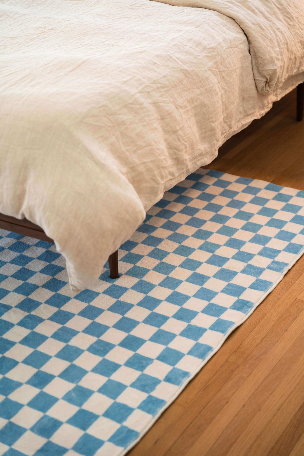 Blue checkered rug partially tucked under a bed, bringing a soft touch of pattern and color to a sunlit minimalist bedroom space.