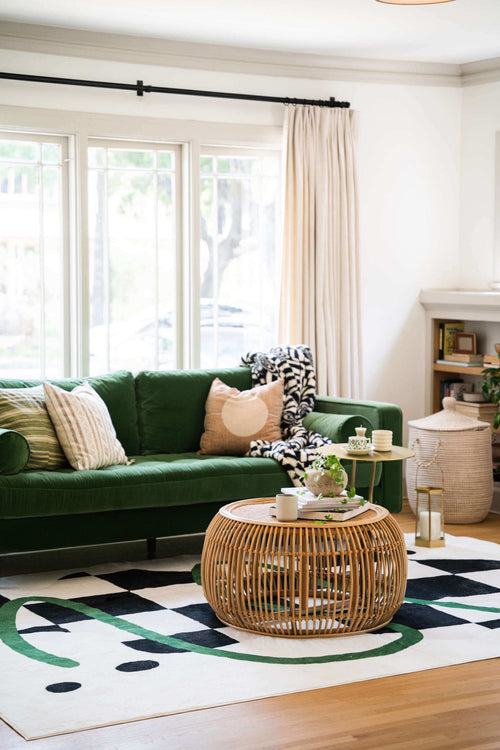 Checkered rug with green swirl under a rattan coffee table, styled with a green velvet sofa and layered cushions.