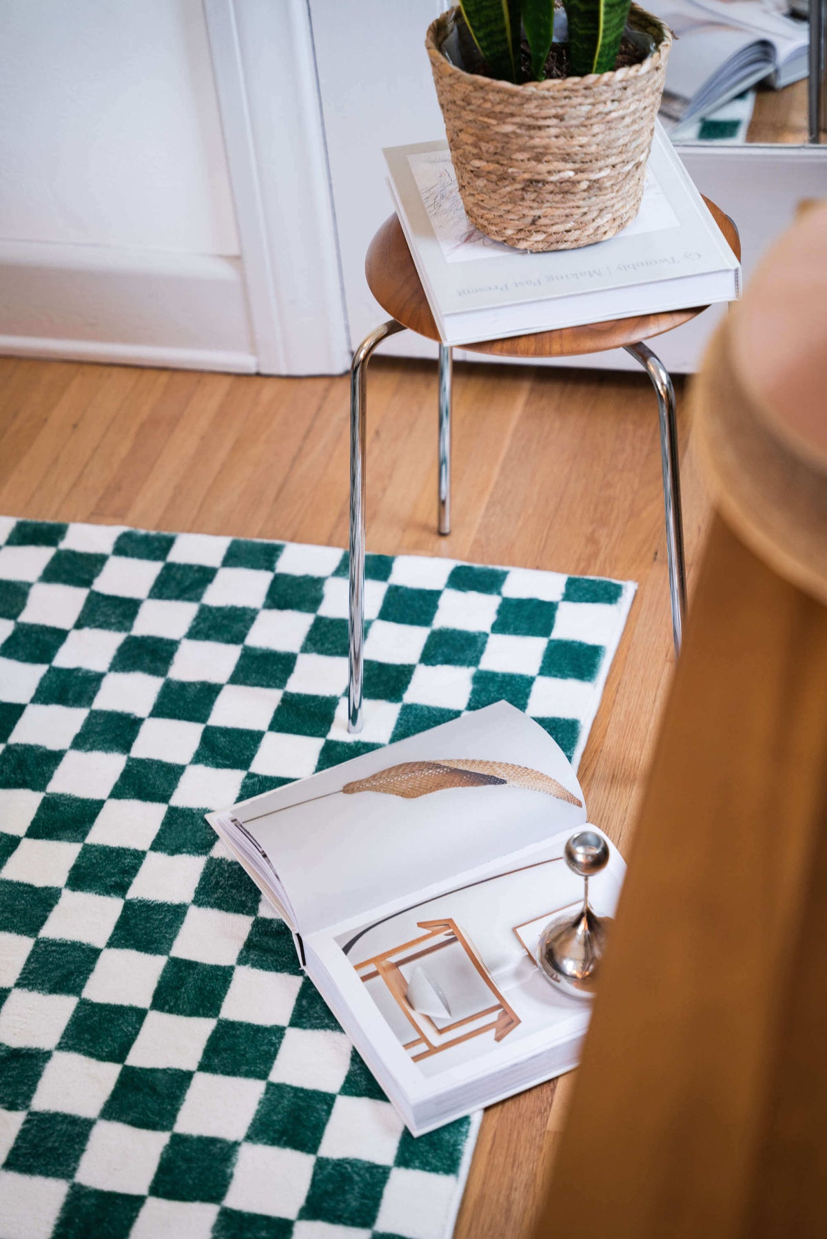 Vibrant checkered rug styled next to a wooden stool stacked with books and a woven planter in a bright indoor setting.