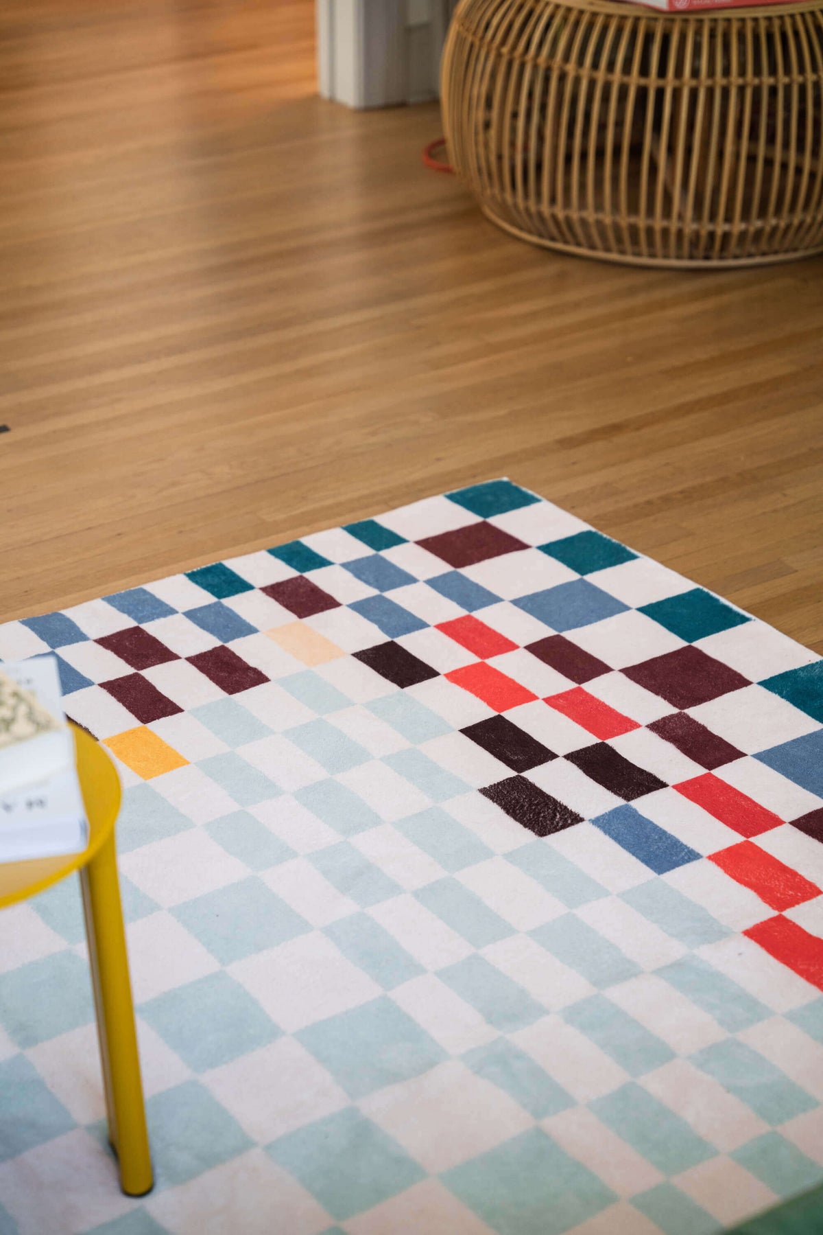 Close-up of the vibrant pixel rug showing red, teal, and maroon checker pattern on pale blue ground near a yellow side table.